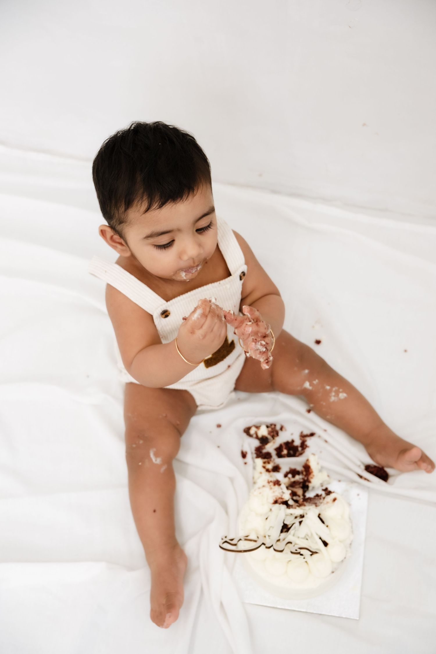 Cute baby in white romper smashing first birthday cake, covered in frosting, sitting on white backdrop.