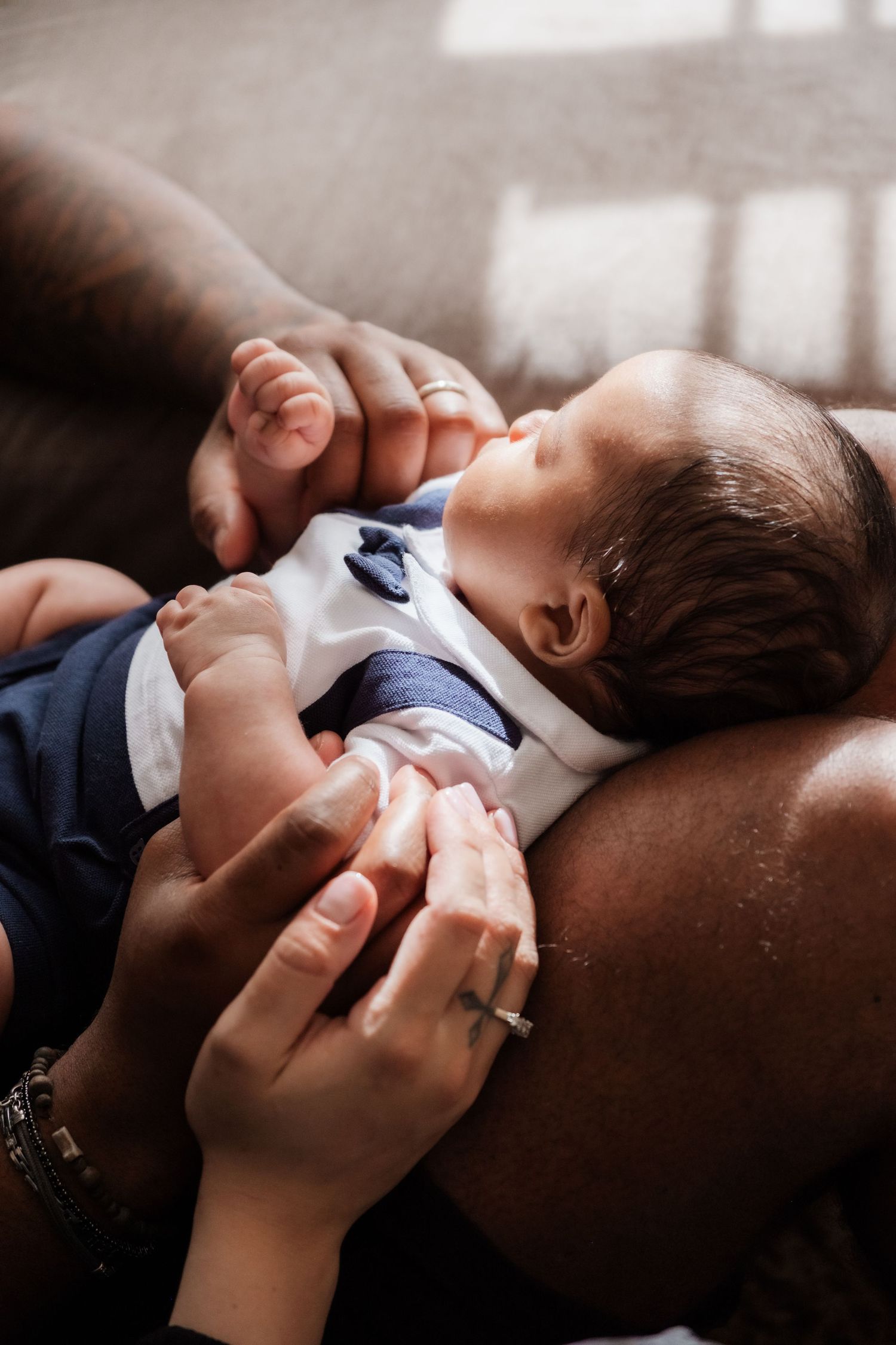 Newborn baby in white and navy outfit resting on parent's chest while both parents gently hold tiny hands.
