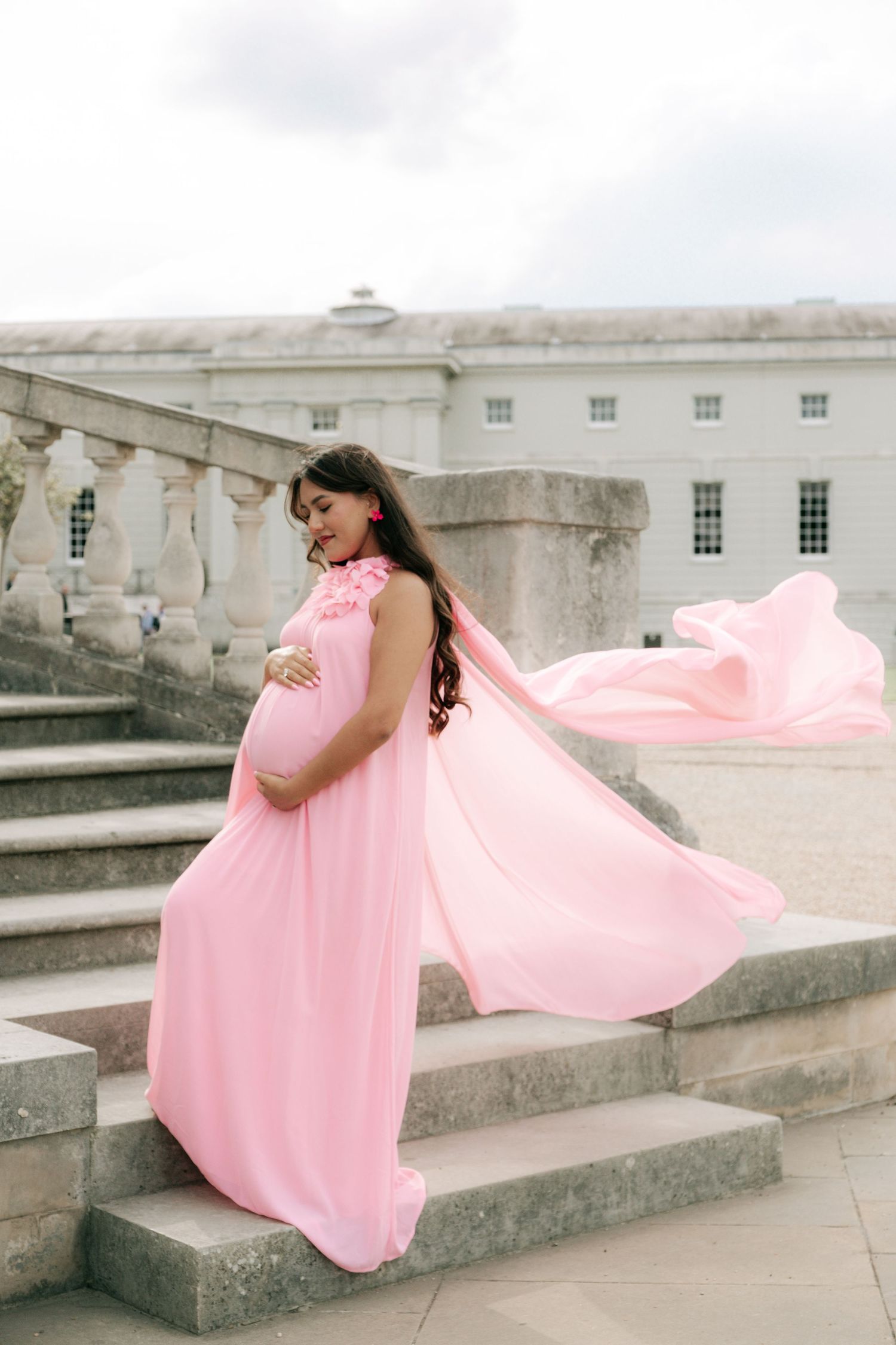 A flowing pink dress billows in the wind as someone poses on stone steps of a historic building.