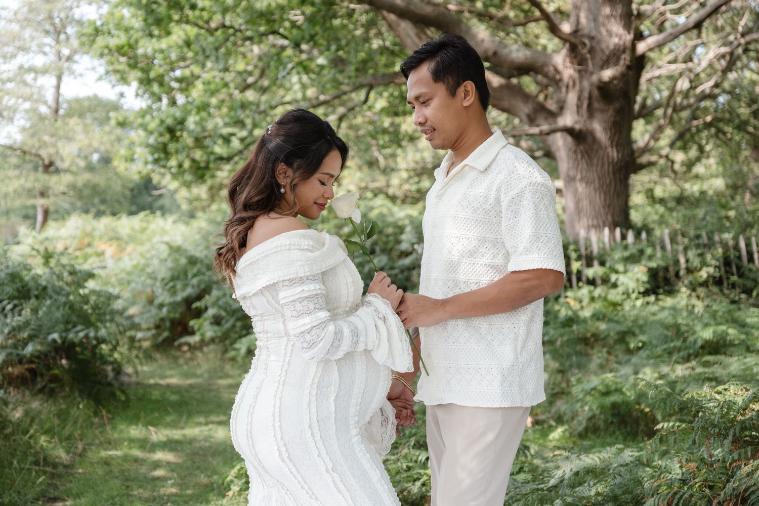 Candid maternity photos of couple in white outfits surrounded by greenery.