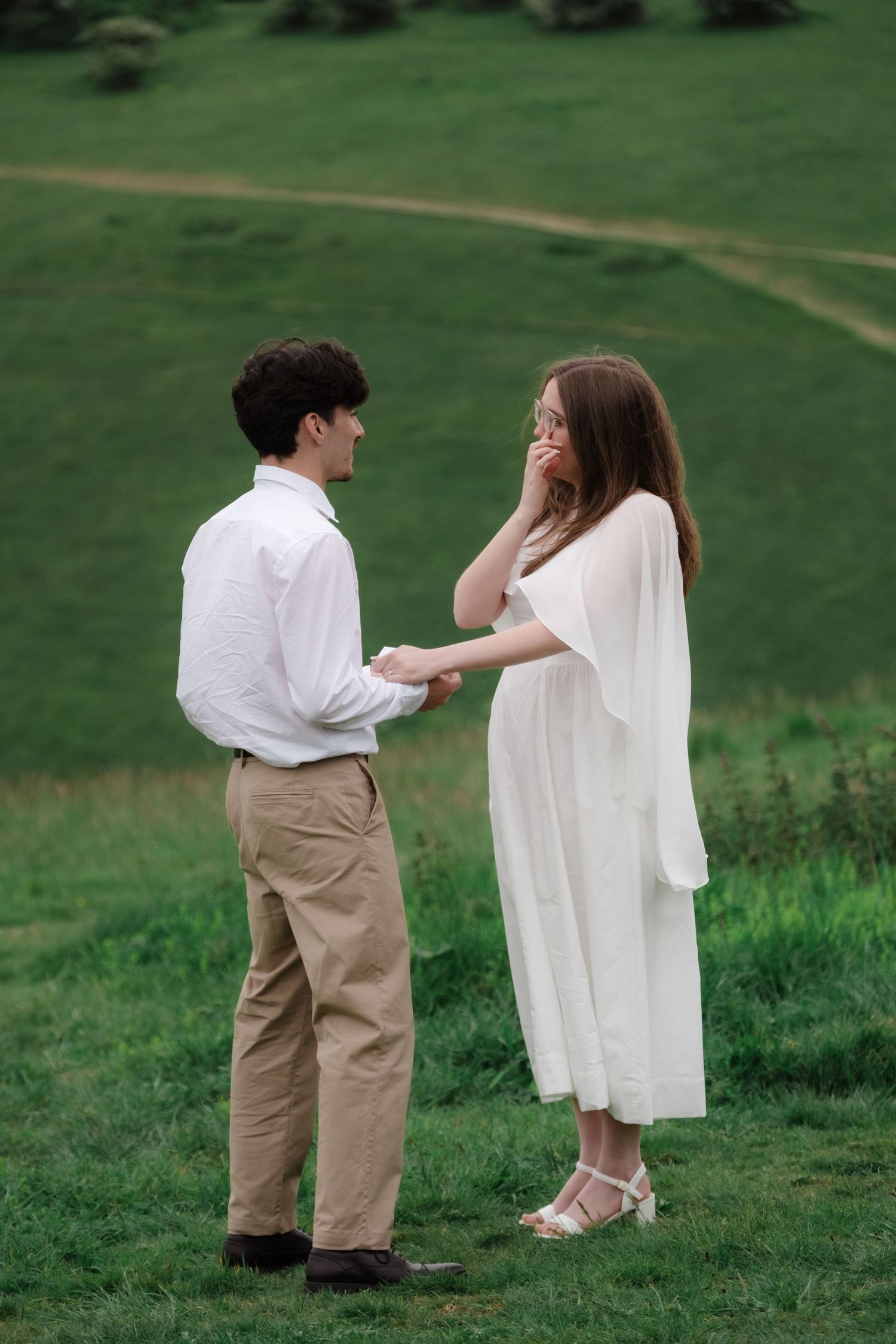 A couple holds hands in a green field, woman in white dress, man in white shirt and khaki pants.