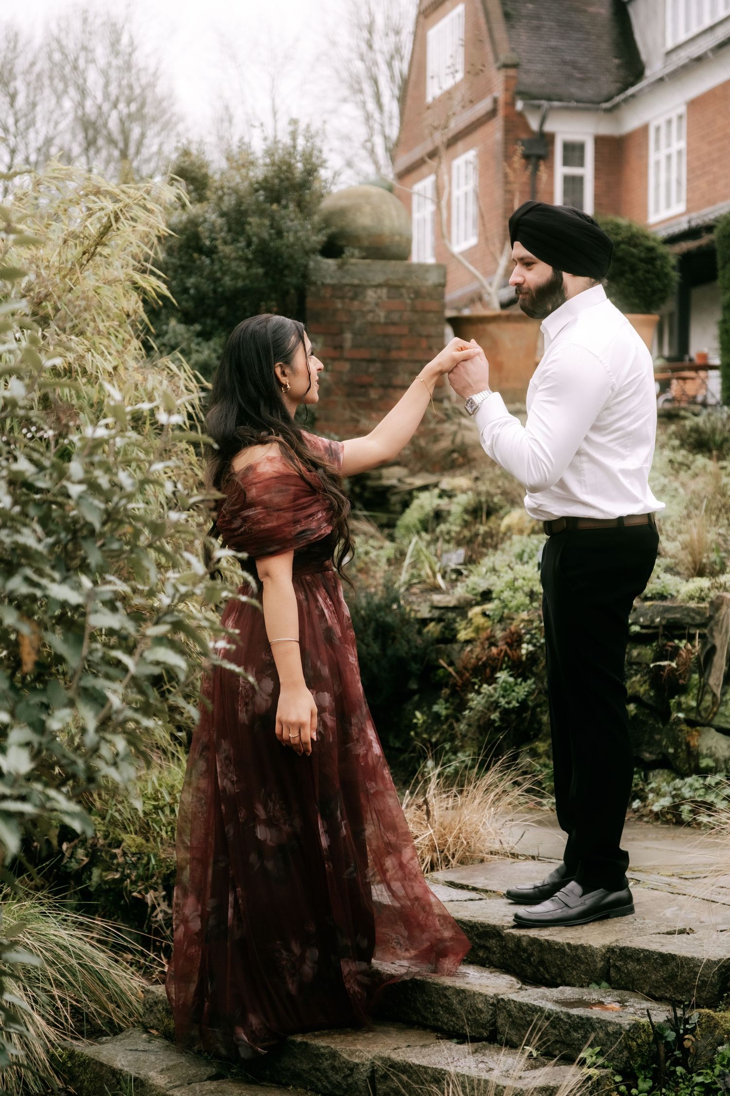 Dancing couple in burgundy dress and white shirt amid garden setting.