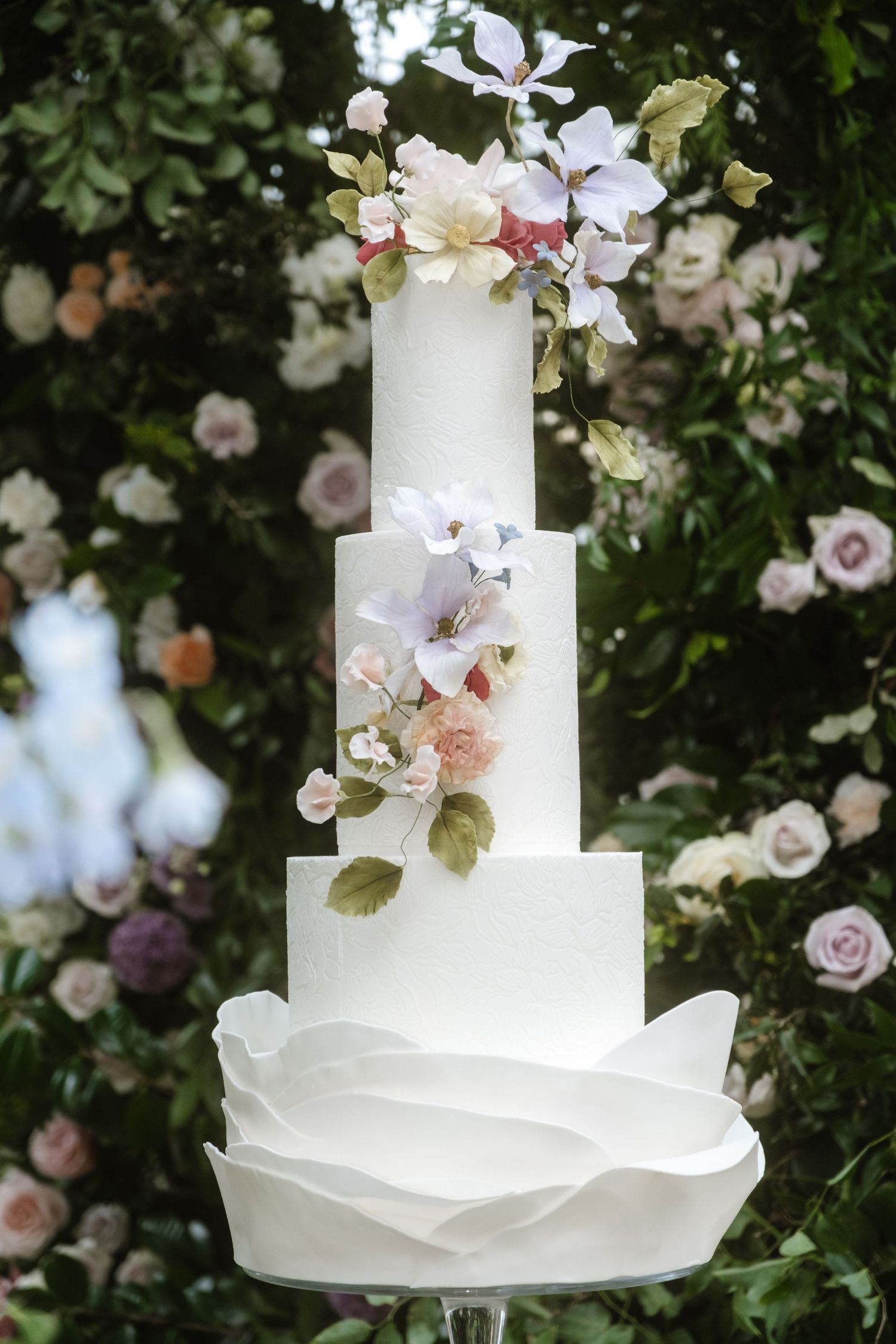 Elegant white wedding cake decorated with delicate pink and white flowers against a garden backdrop.