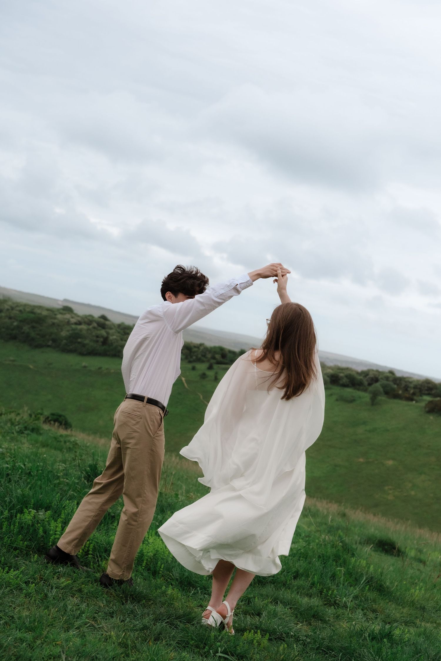 Couple dancing in a green field, woman in flowing white dress, man in white shirt and khaki pants, cloudy sky backdrop.