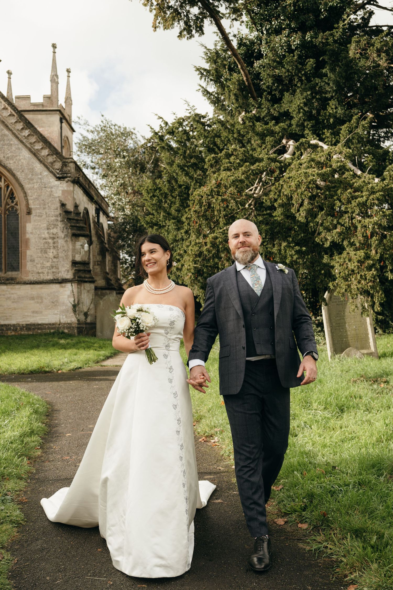 Newlyweds walk together along church pathway surrounded by greenery.