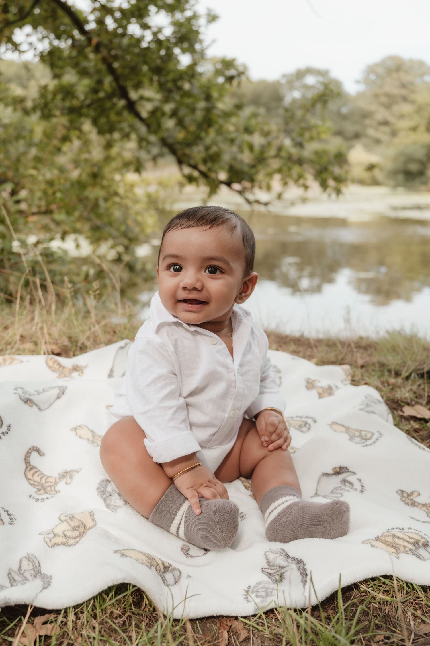 Baby in white clothing lies on a patterned blanket outdoors near a pond.