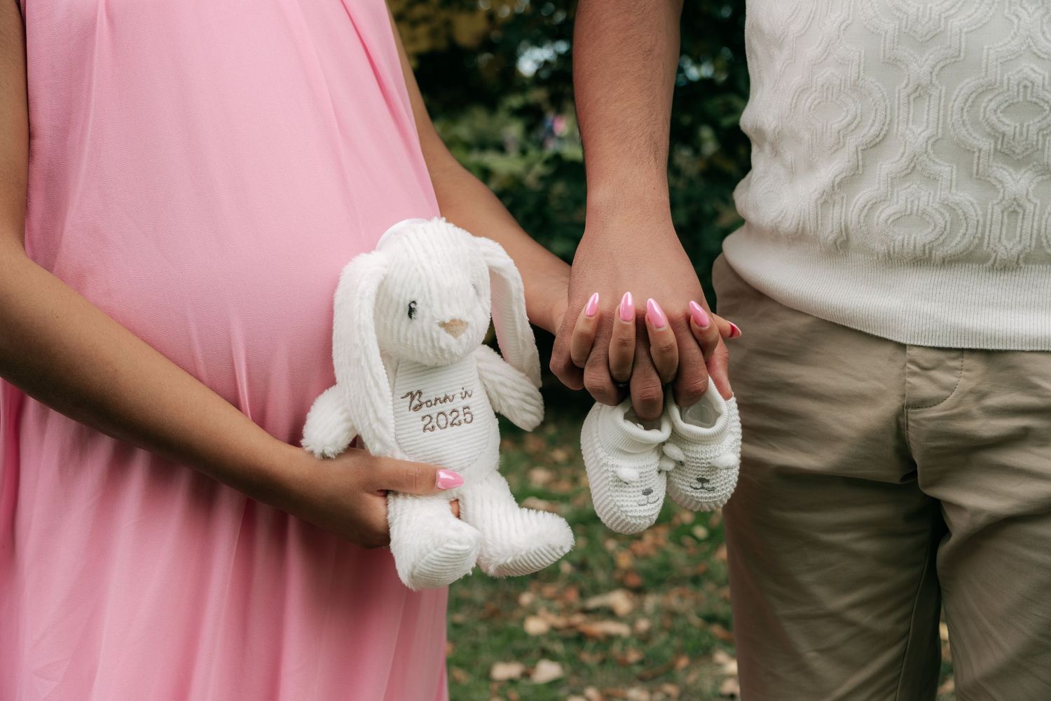 Hands hold tiny white baby shoes against a pink and beige color palette.