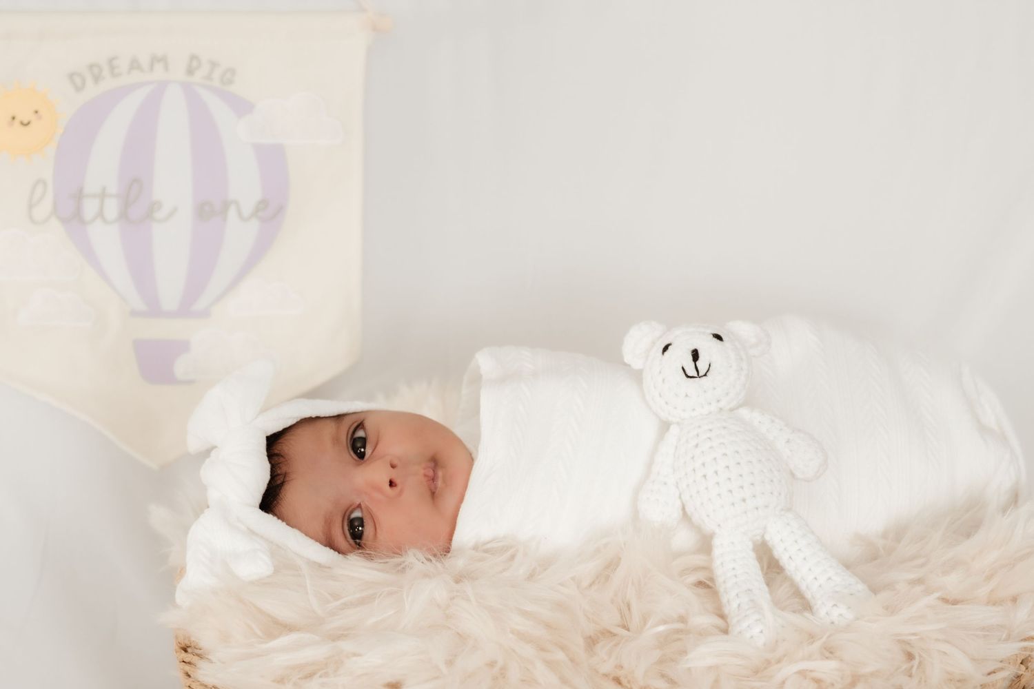 Newborn baby wearing a white knit bear hat rests peacefully on white bedding.