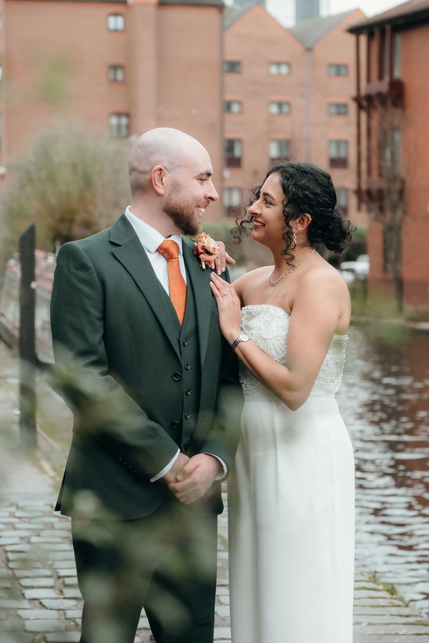 A couple shares an intimate moment on their wedding day by a waterfront with brick buildings in the background.