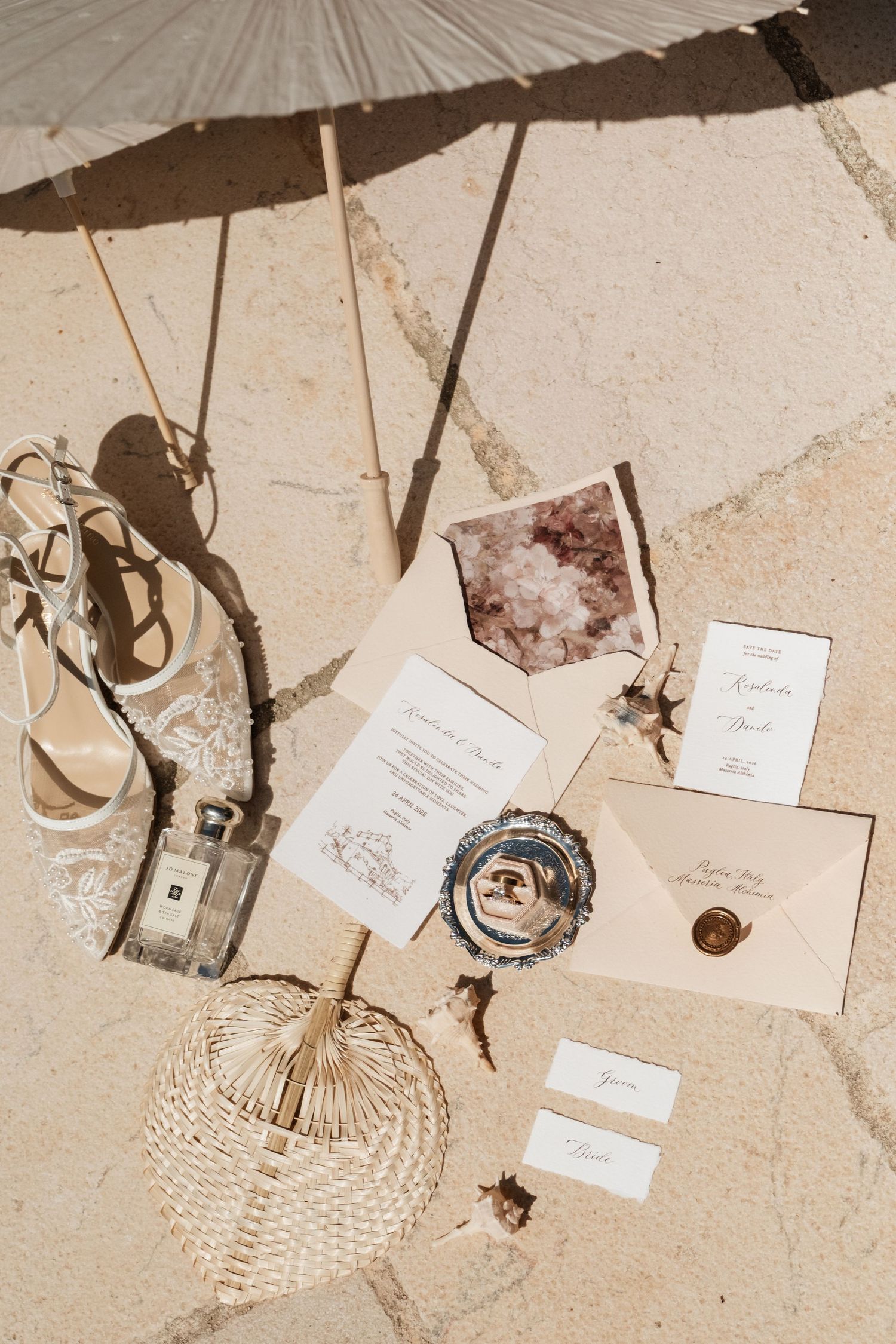 Elegant wedding flat lay with invitations, rings, lace shoes, perfume, and parasol on stone surface.