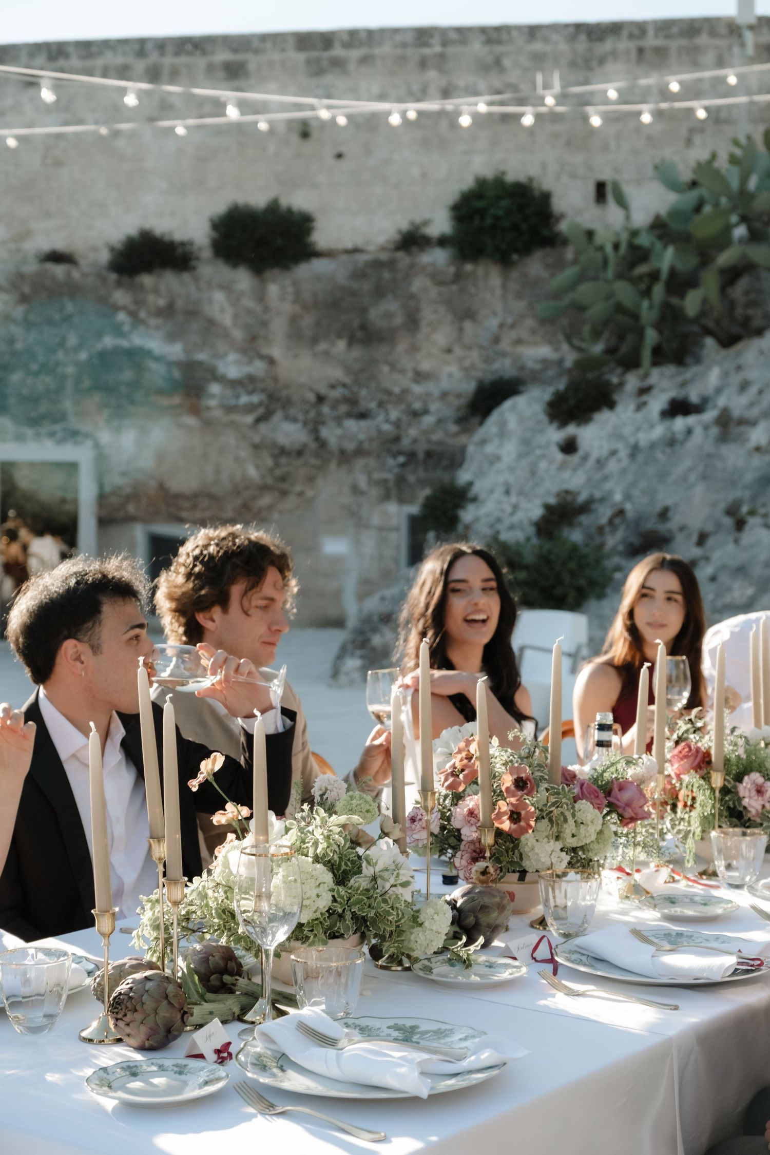 Wedding guests laugh and celebrate at an elegantly decorated outdoor reception table with floral arrangements and candles.