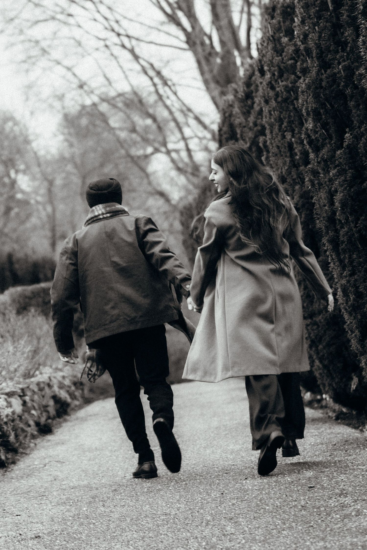 A couple runs together holding hands along a garden path in winter coats.