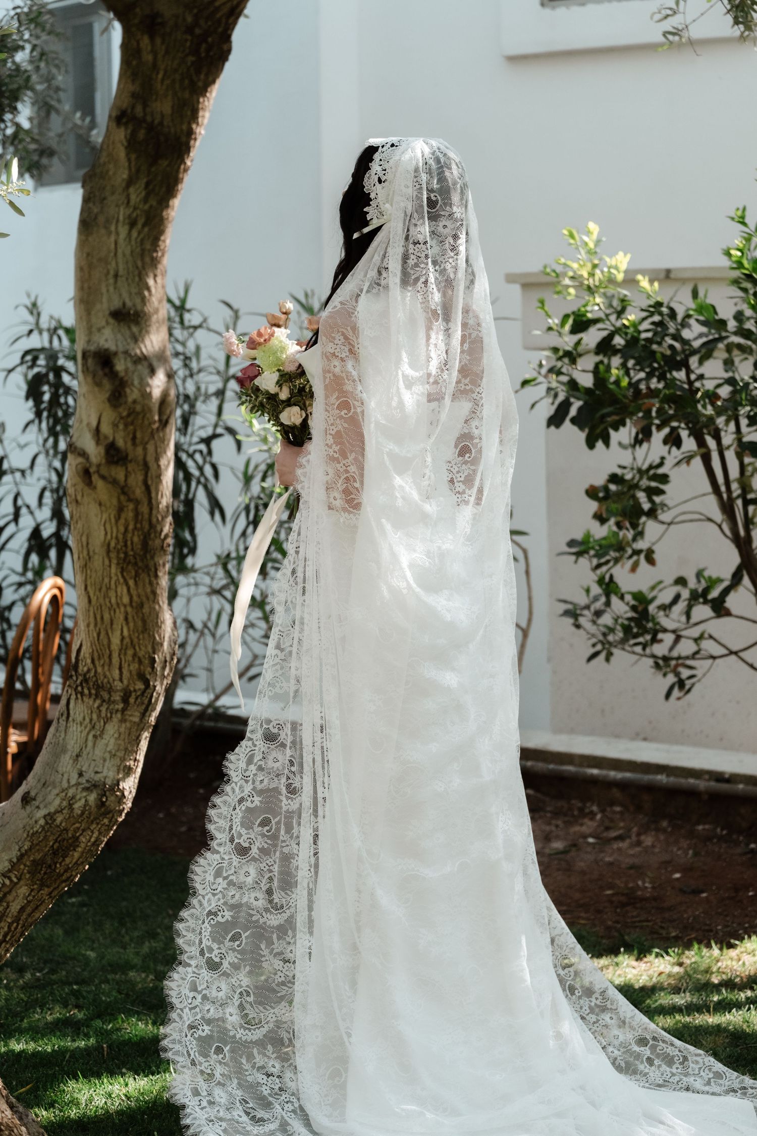 Bride in elegant lace wedding gown with long cathedral veil holds bouquet in sunlit garden setting.