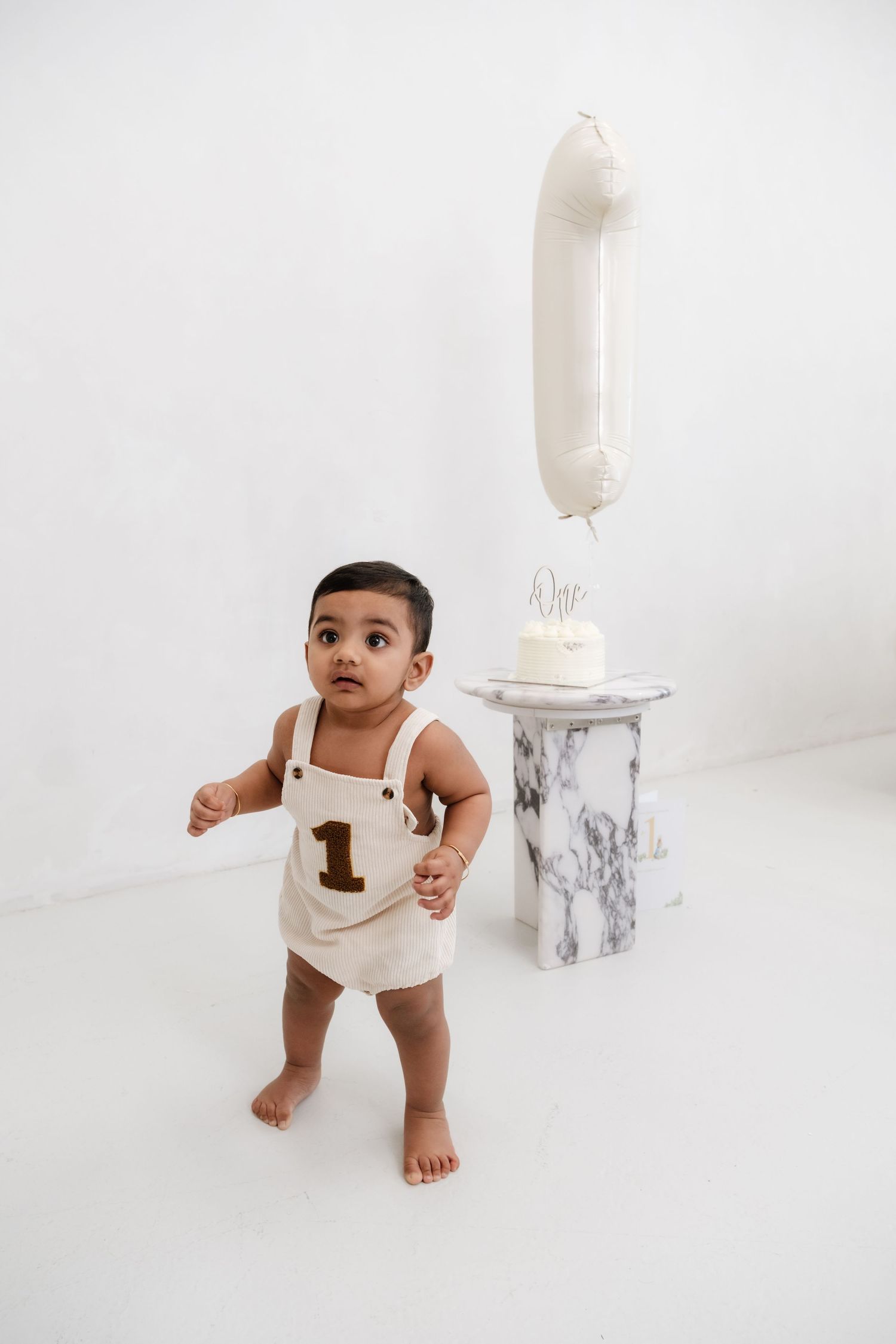 A toddler in a cream romper with number 1 stands near a marble pedestal with a white birthday cake and balloon.