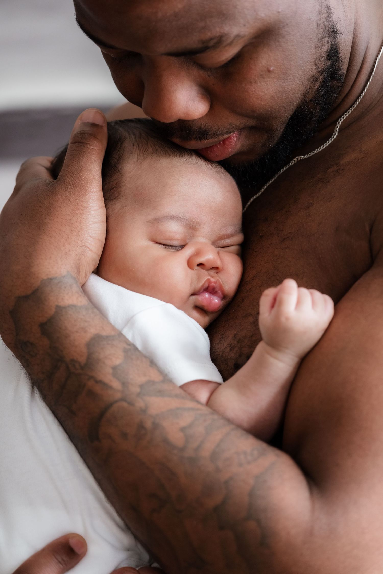 A shirtless tattooed father tenderly holds his sleeping newborn baby close to his chest.
