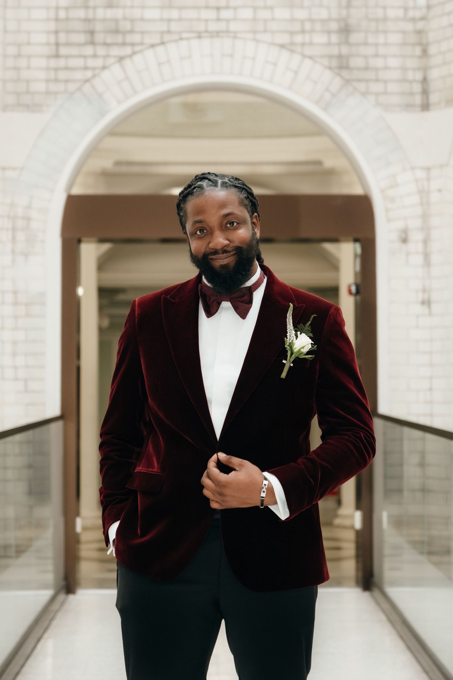 A person in a burgundy velvet tuxedo jacket poses in an elegant white brick hallway.