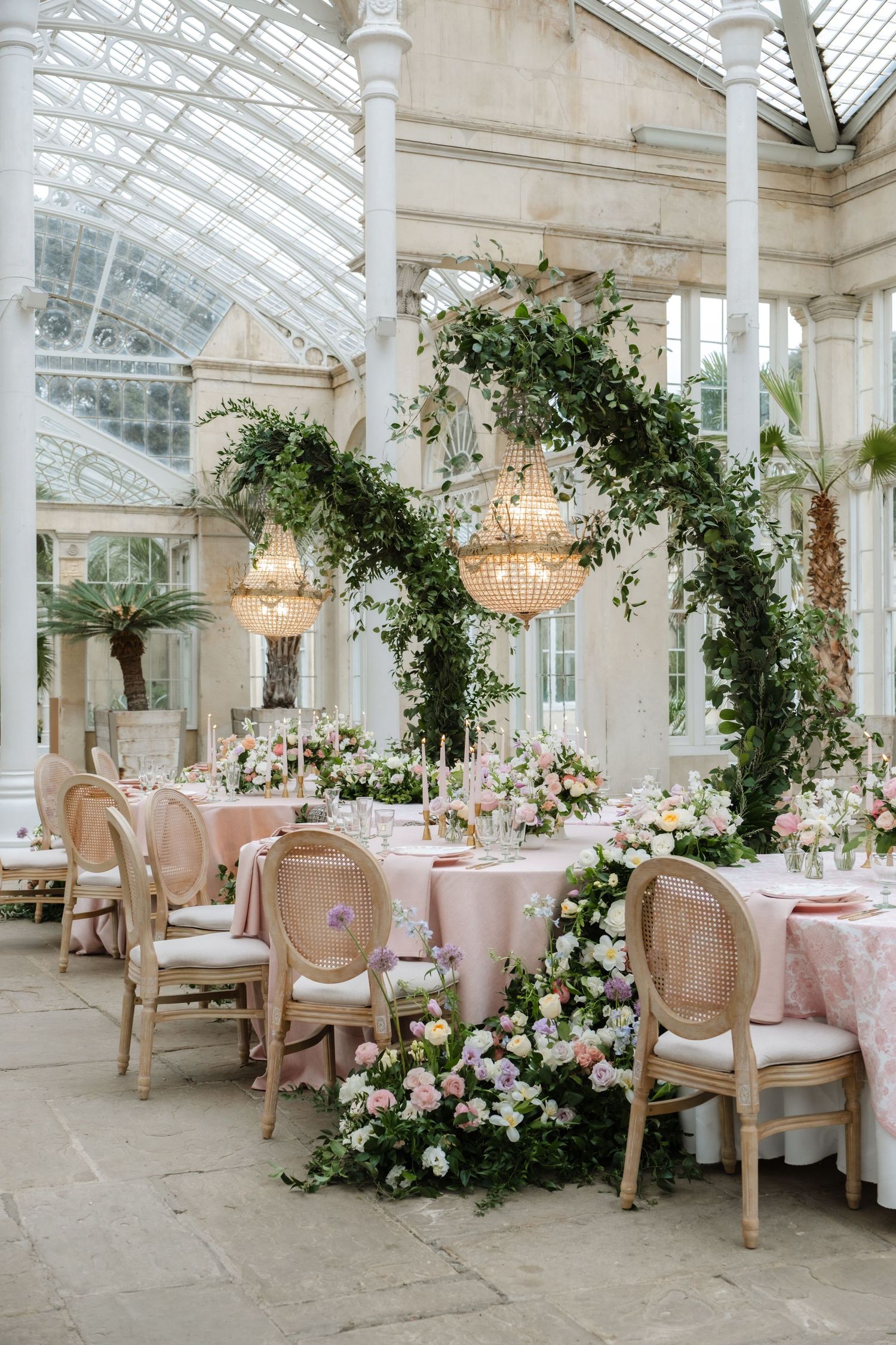 Elegant conservatory reception space with pink tablecloths and floral arrangements under glass ceiling.