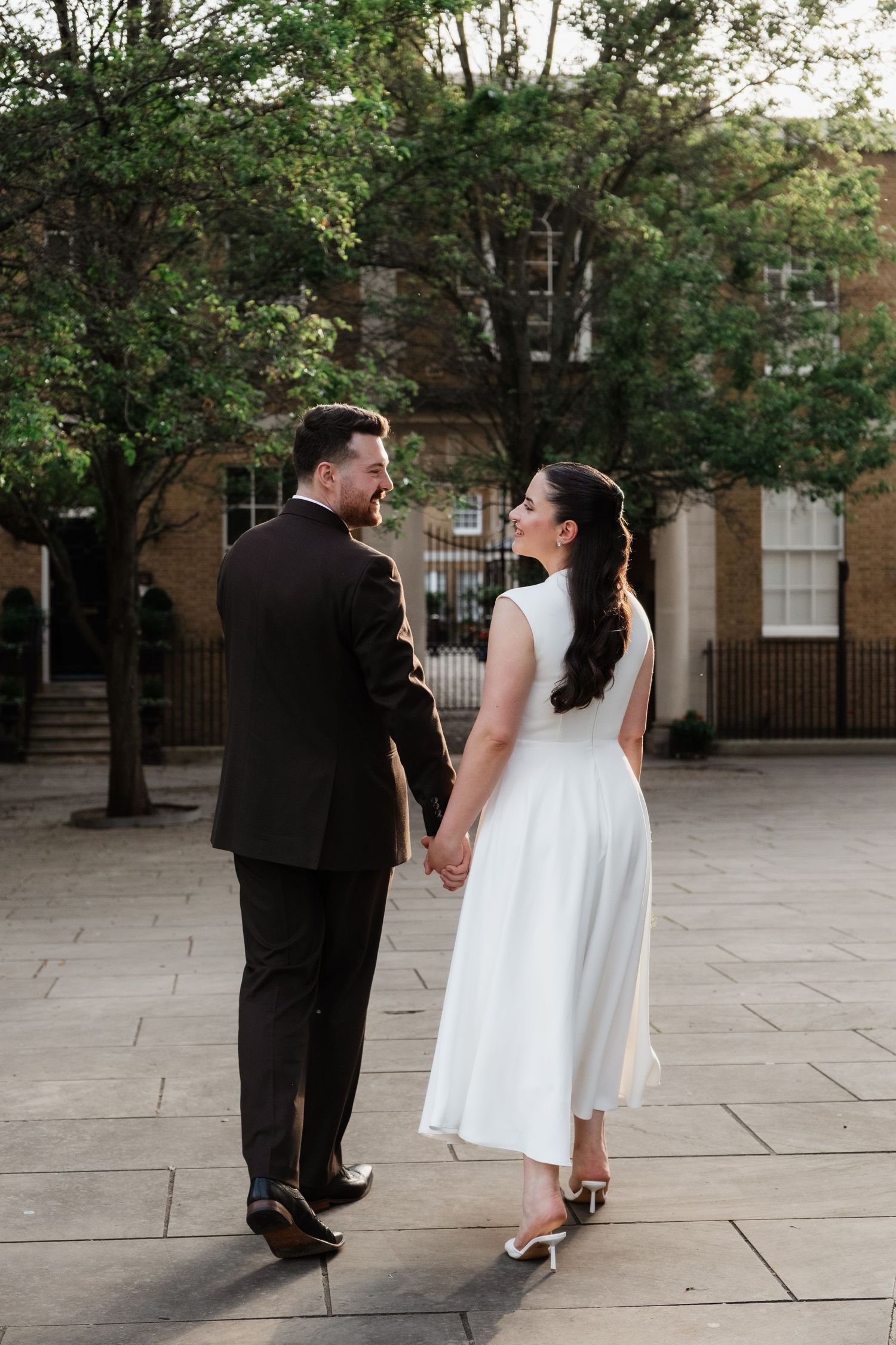 A bride and groom walk hand-in-hand in a courtyard, she in a white midi dress, he in a dark suit, surrounded by green trees.