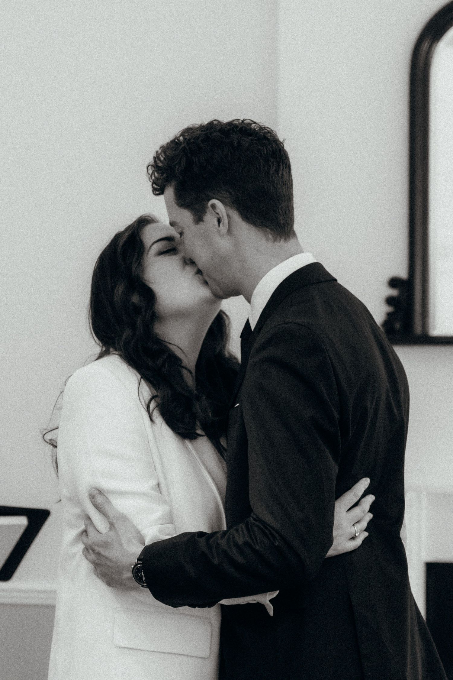 Black and white photo of a couple sharing a kiss, dressed in formal wedding attire, embracing tenderly.