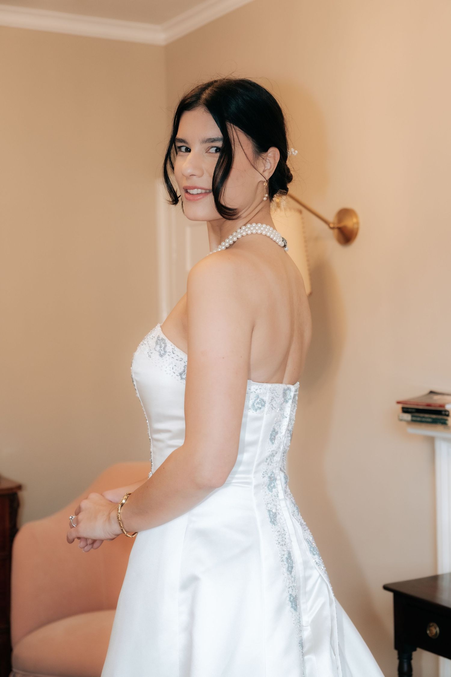 A bride in a white strapless gown and pearl necklace poses elegantly in an indoor setting.