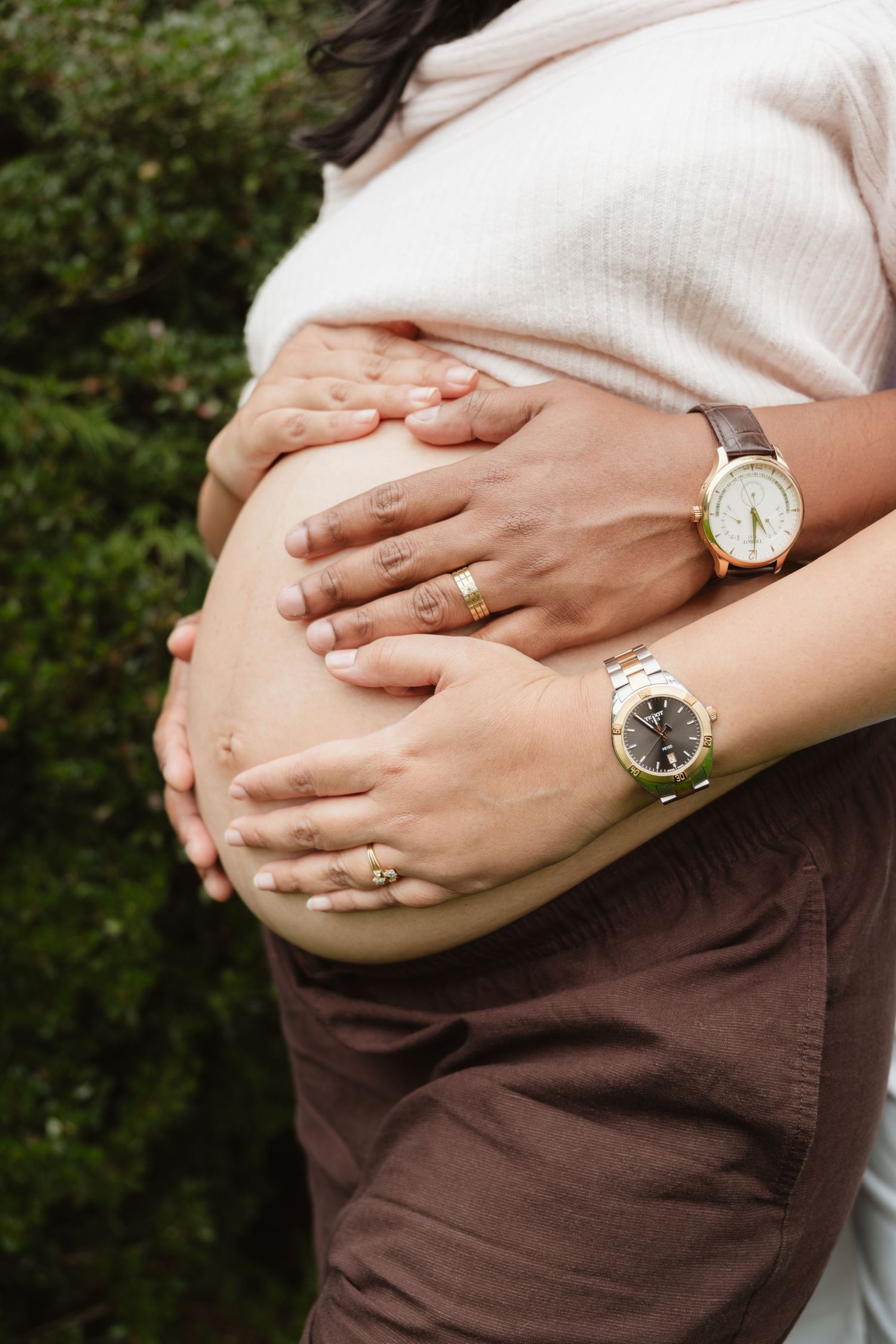 Close-up of hands cradling a pregnant belly, with a gold watch and wedding ring visible.
