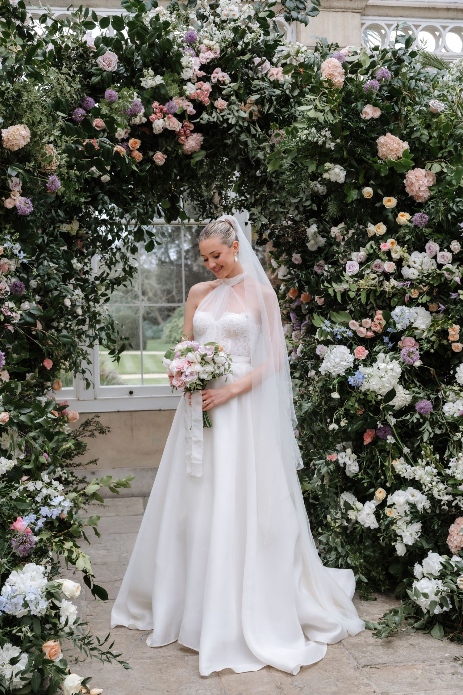 Bride in flowing white gown poses among climbing roses in conservatory setting.