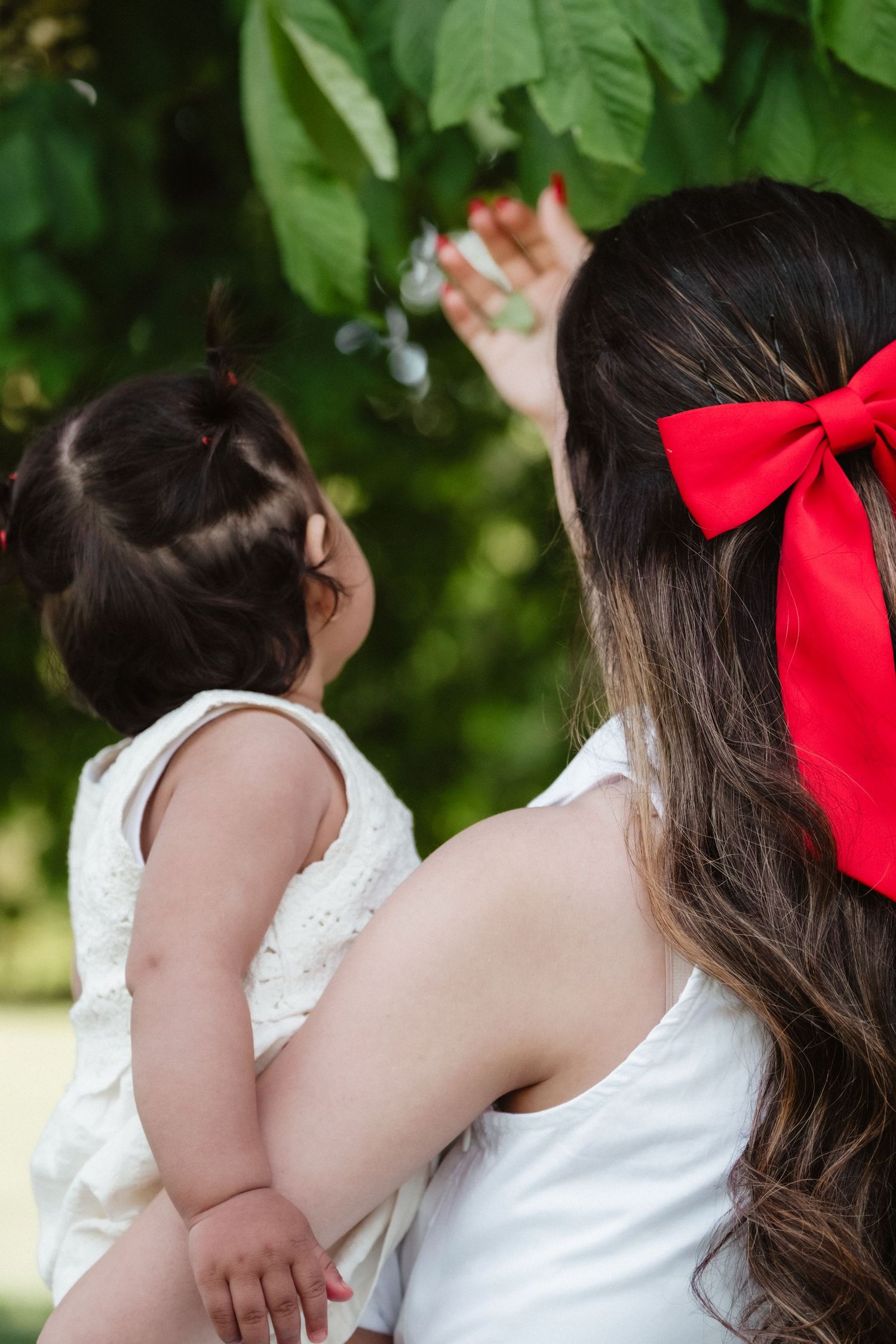 A mother with a red bow holds her baby girl outdoors, pointing at green leaves in a sunlit park.