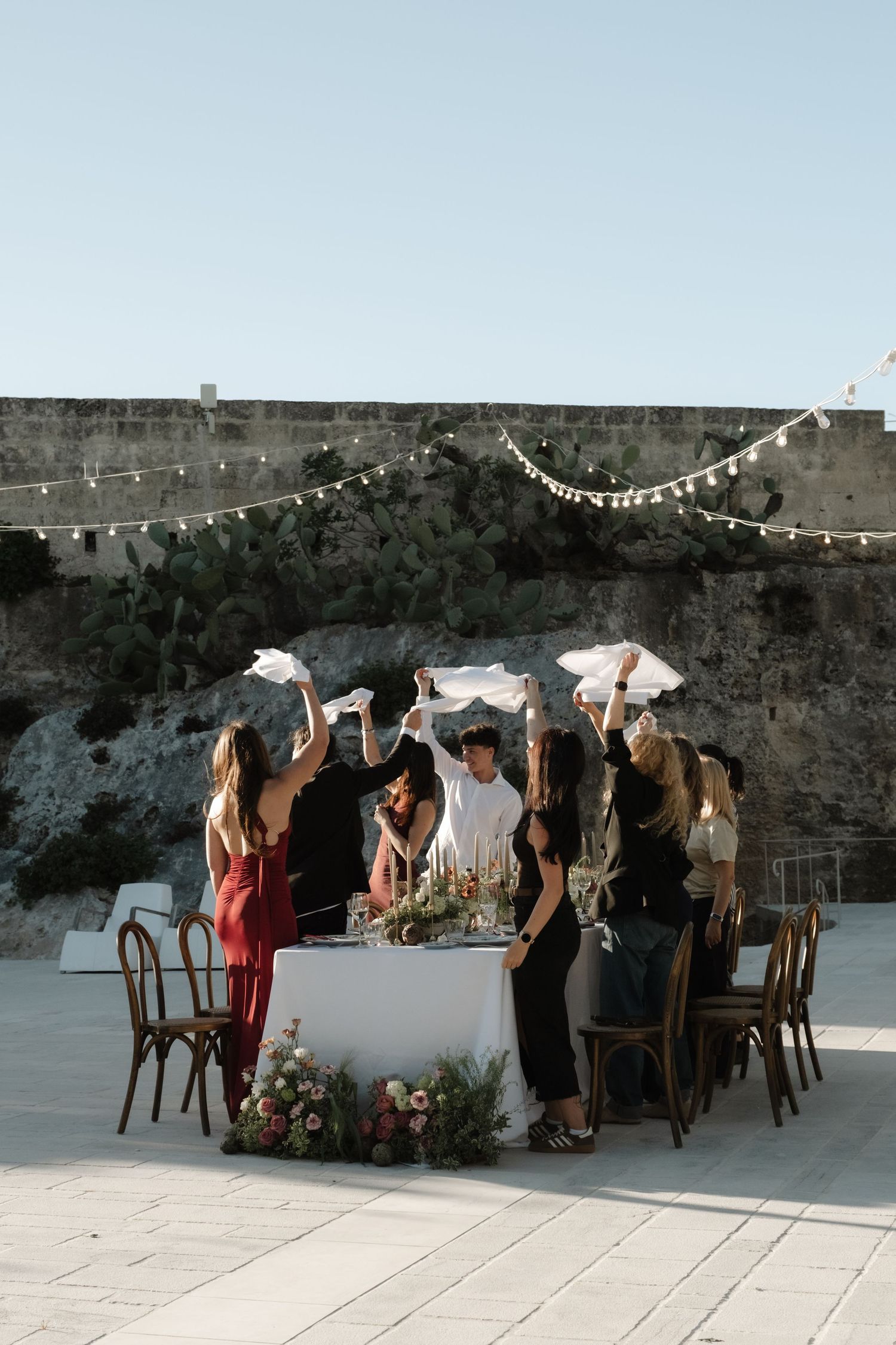 Group of women celebrating at an elegant outdoor wedding table with string lights and stone wall backdrop.