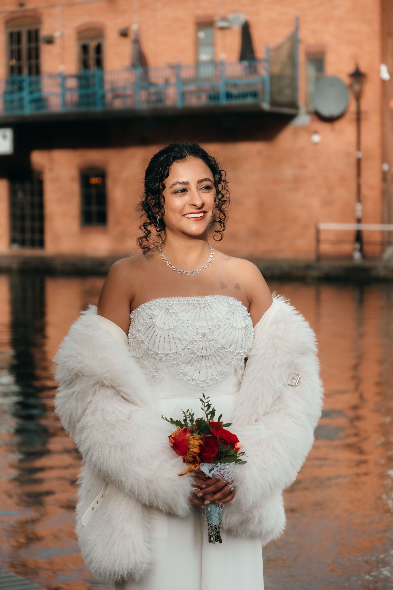 A bride in a white dress and fur wrap holds a red bouquet against a waterfront backdrop.