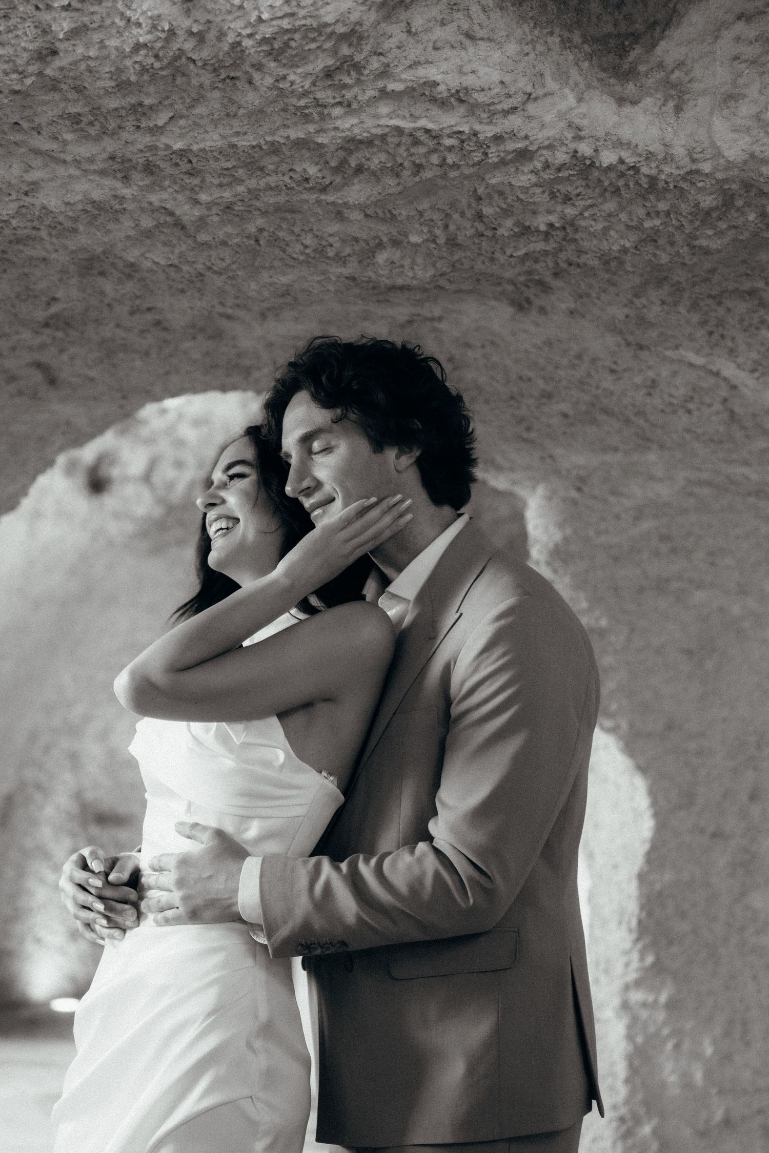 Black and white photo of a couple embracing in a cave, bride in white dress and groom in suit smiling.