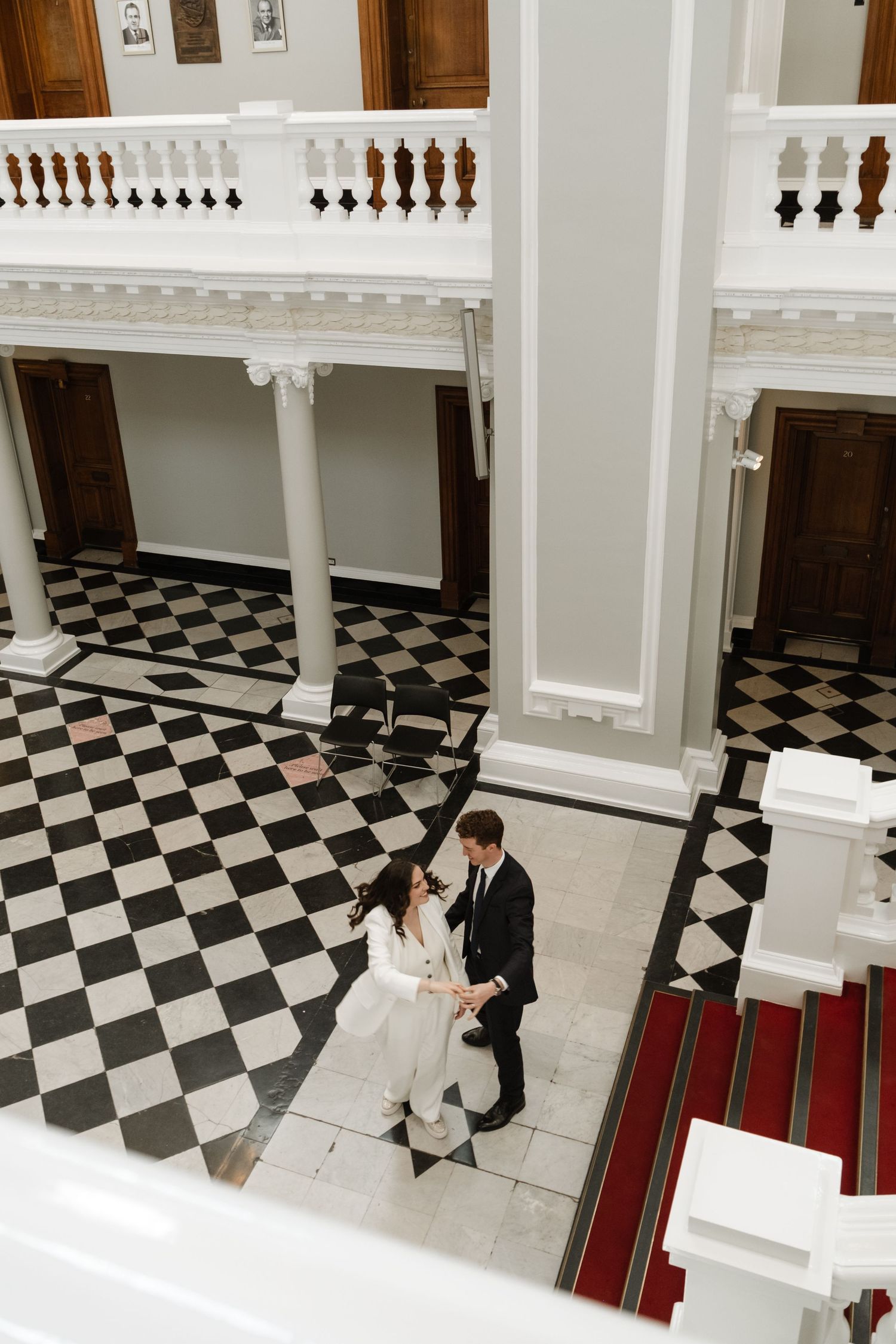 Bride and groom dancing in elegant hall with checkered floors, white columns, and balcony in grand historic venue.