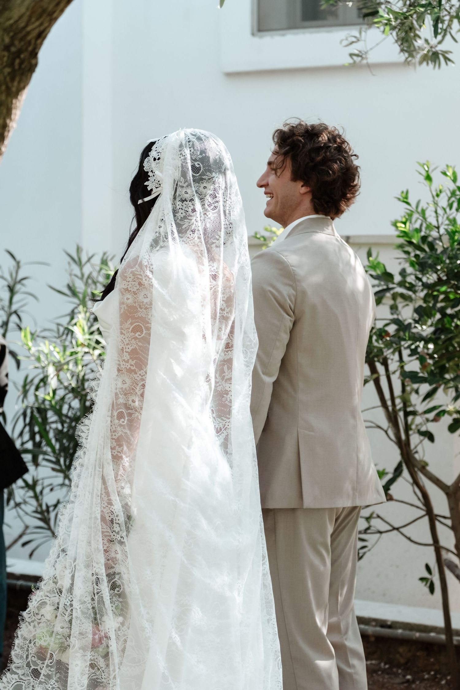 Bride in lace veil and groom in beige suit stand together outdoors, smiling, with greenery and white building behind them.