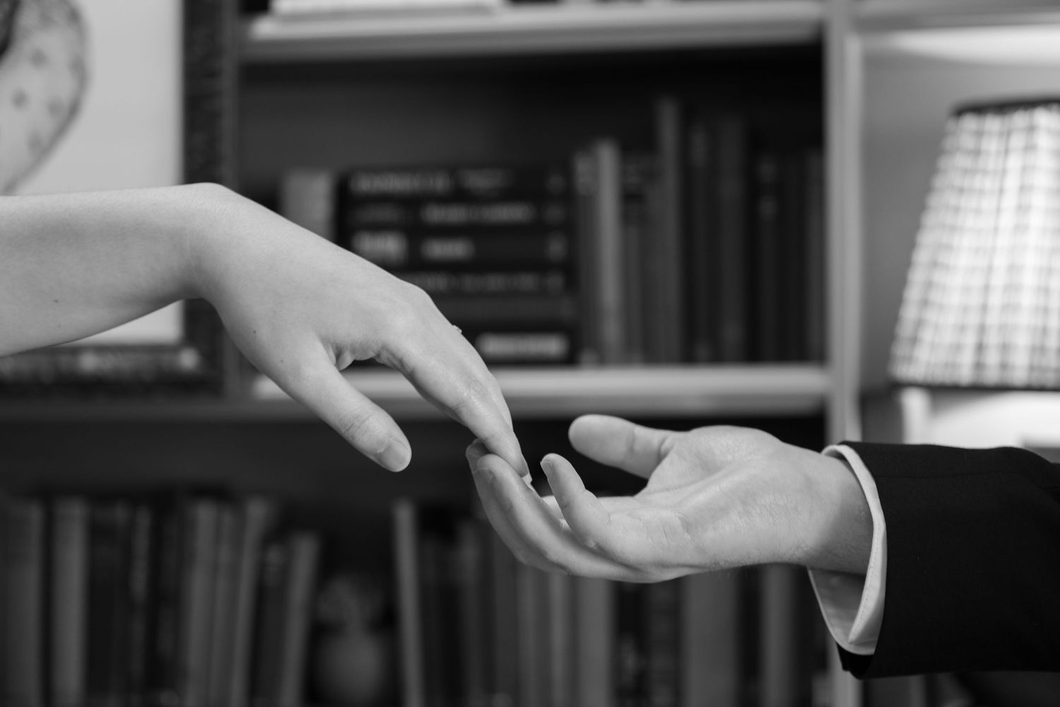Black and white photo of two hands gently reaching toward each other in front of a bookshelf.