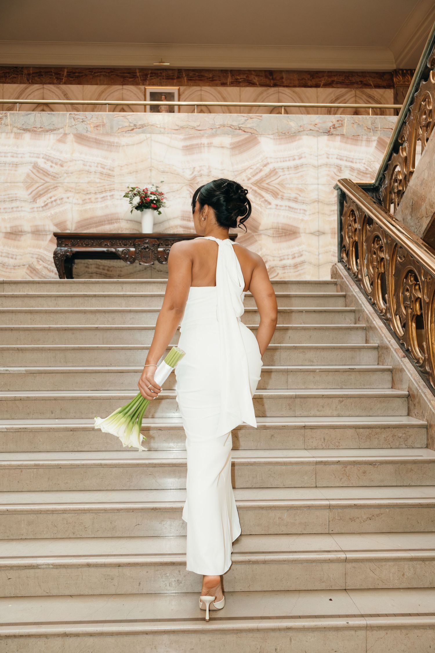 Bride in sleek white dress ascending marble staircase while holding bouquet.