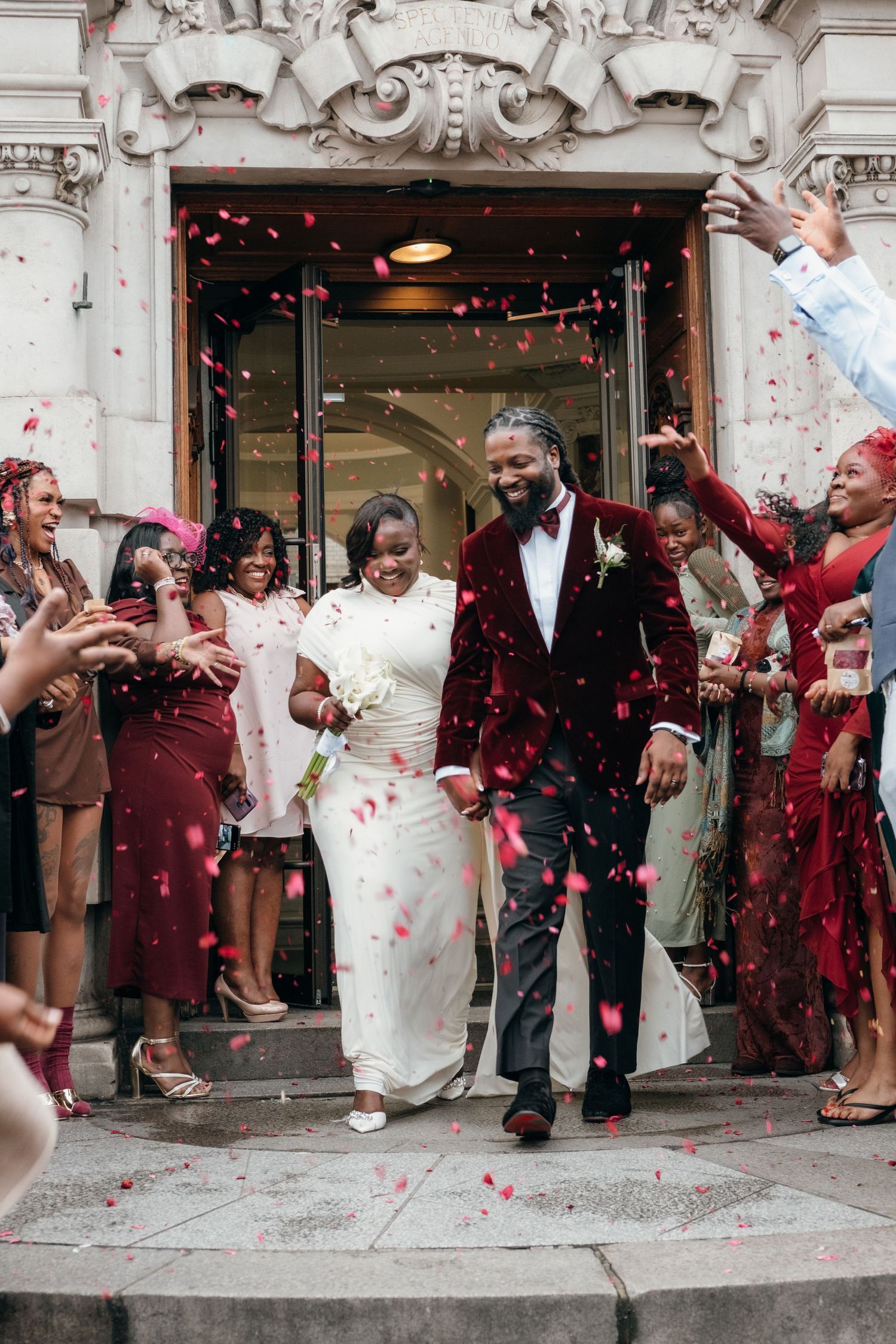 Wedding party exits building as guests shower them with red rose petals in celebration.
