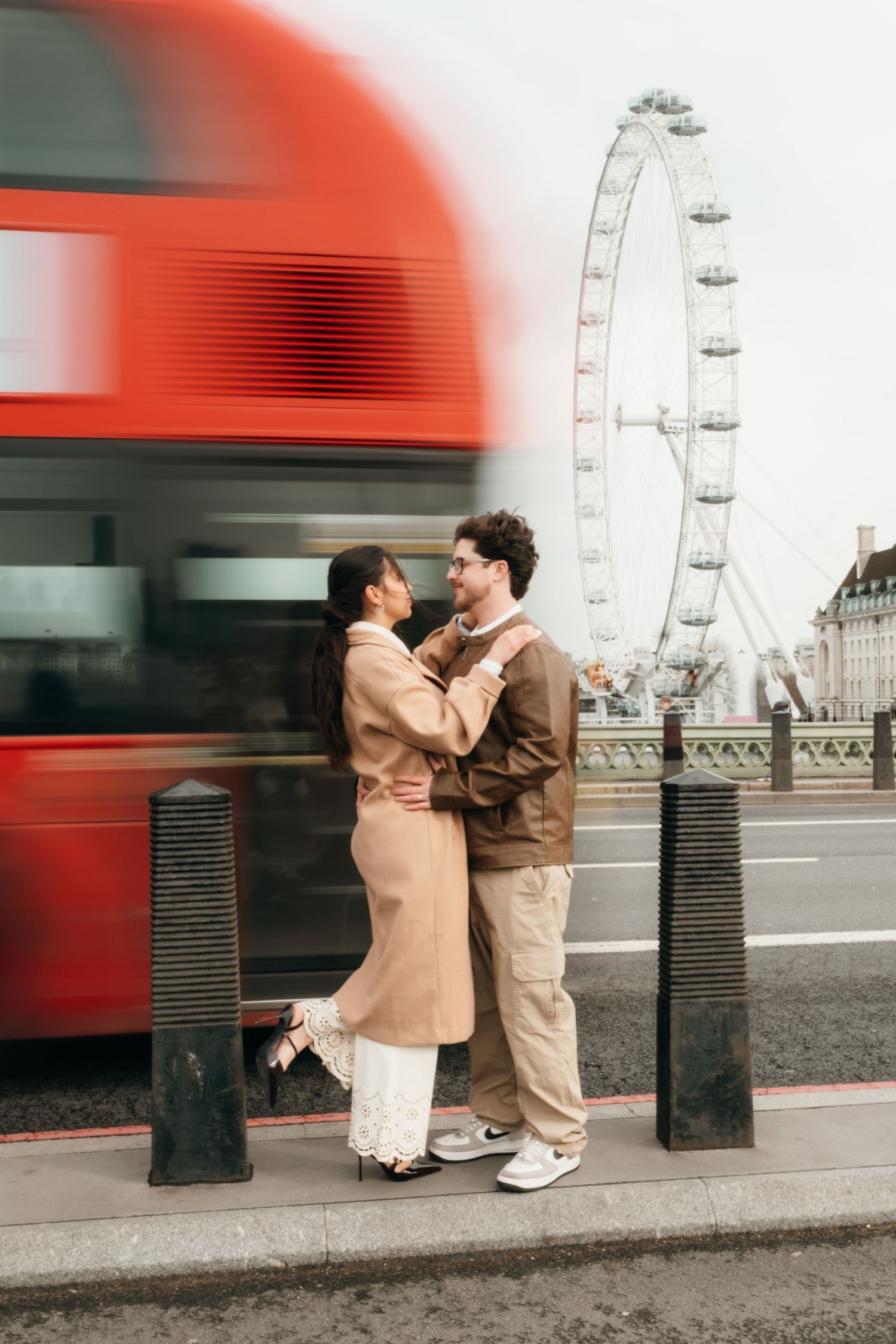 Couple embracing on Westminster Bridge with the London Eye and a blurred red double-decker bus in the background.
