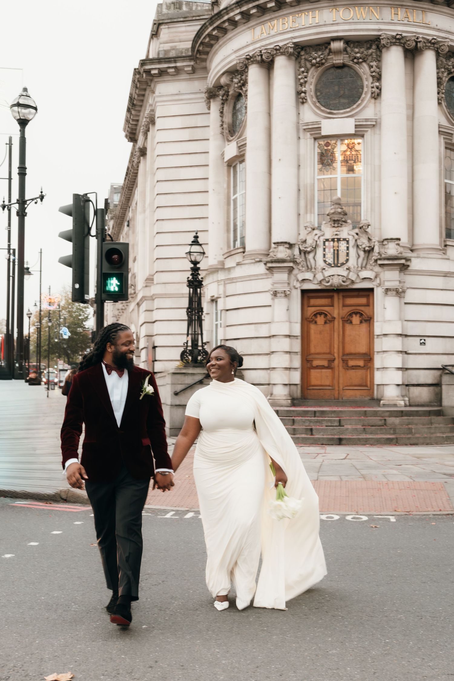 A couple in wedding attire walks hand in hand on a city street in front of an ornate historic building.
