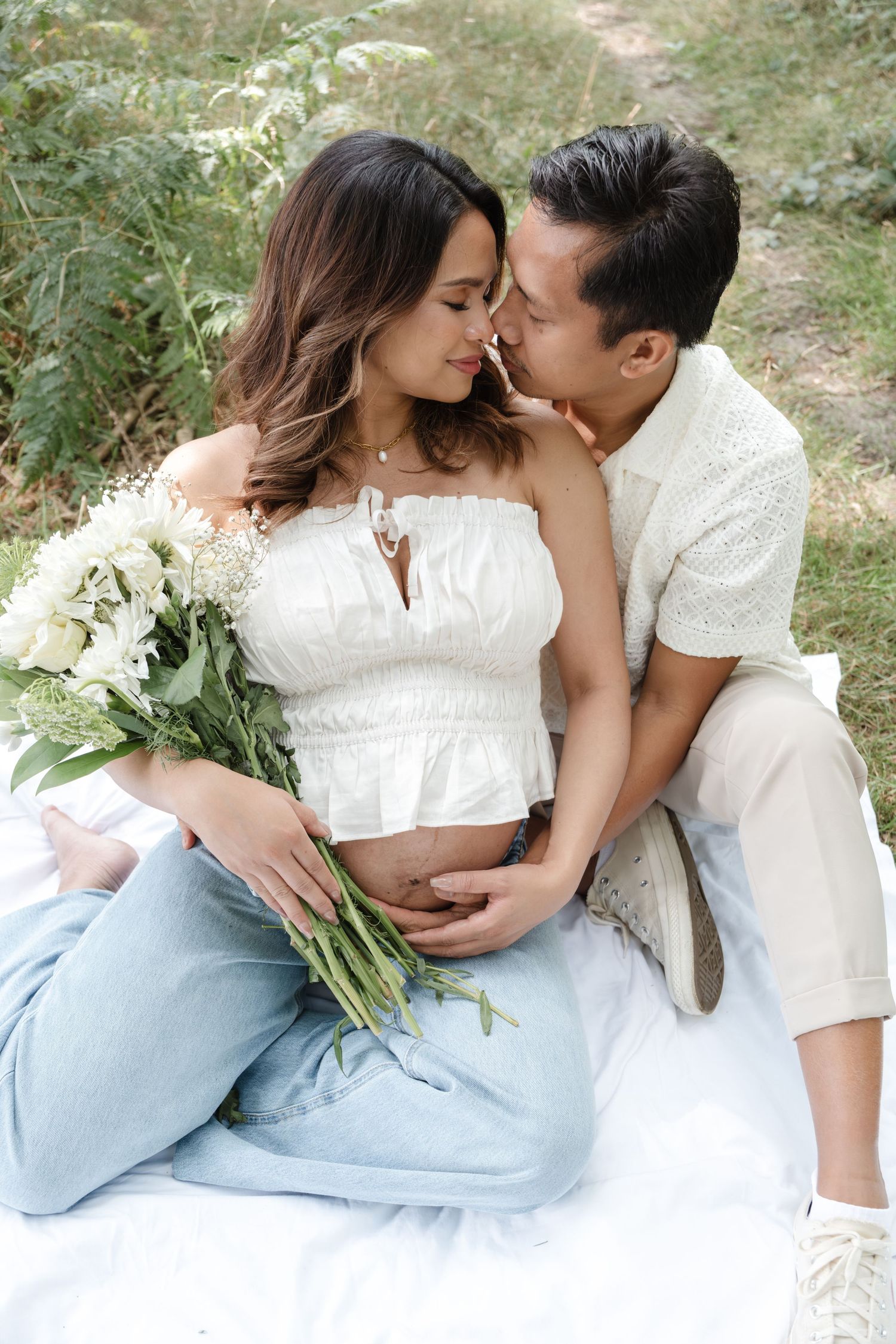 Romantic couple in white outfits share tender moments while sitting together outdoors with greenery in background.