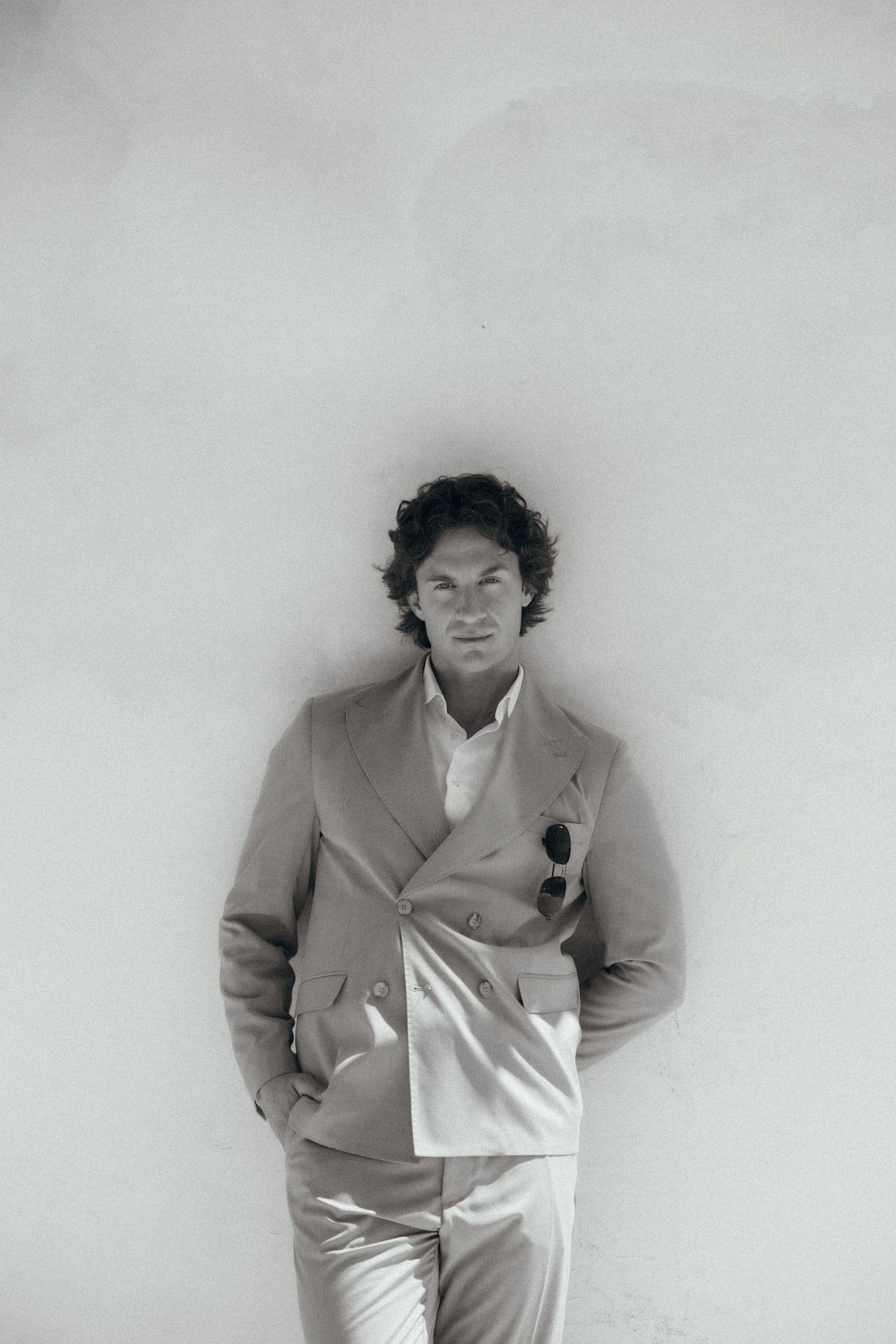 Black and white portrait of a stylish man in a double-breasted suit leaning against a white wall.