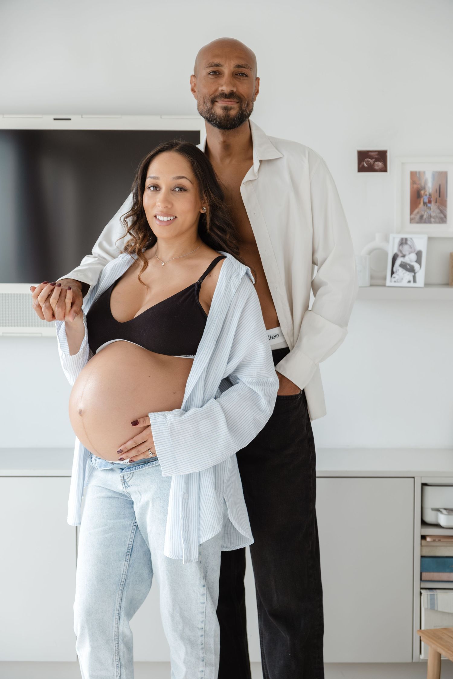 Couple embraces in a bright white modern room during a maternity photoshoot.