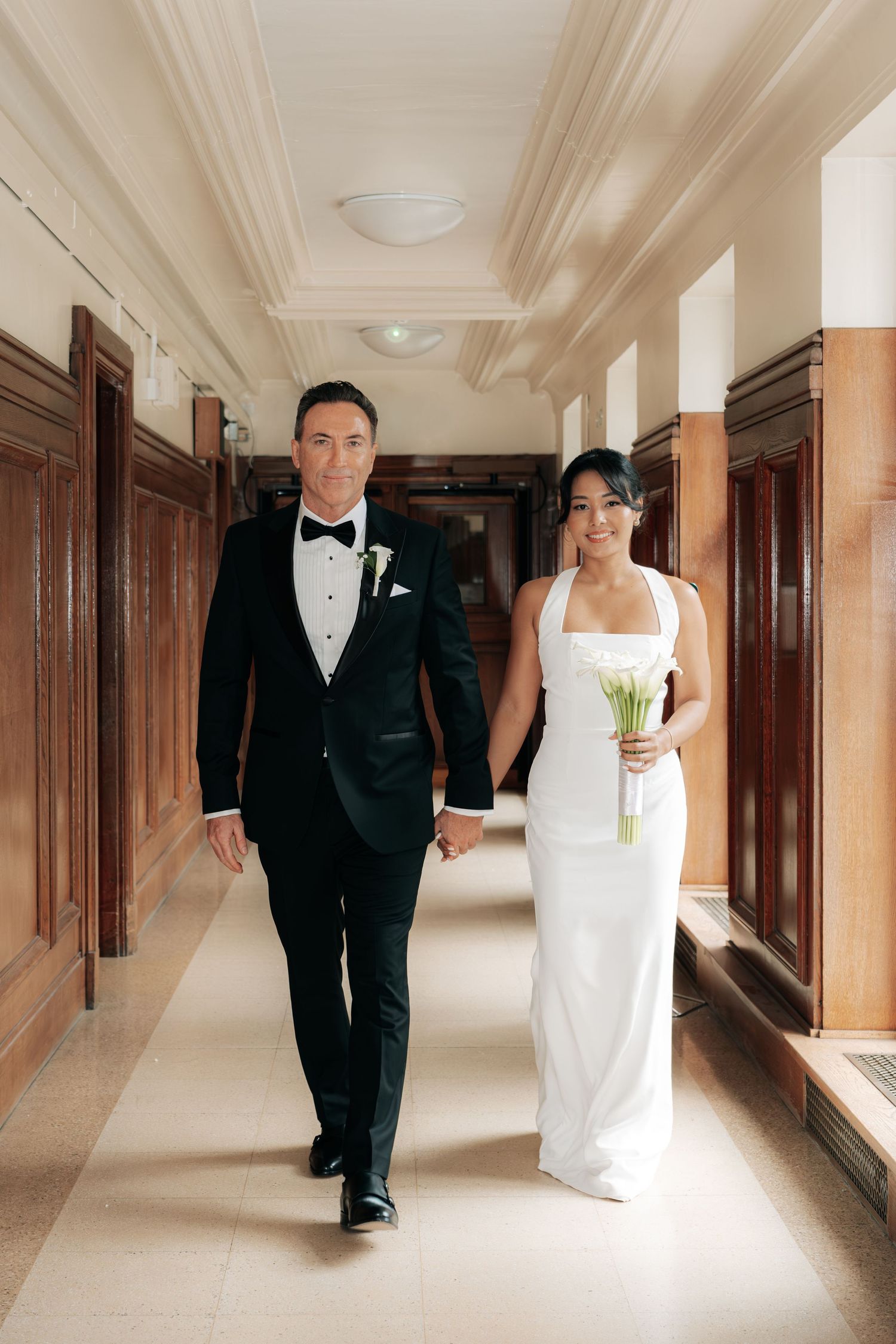 A couple in formal wedding attire walks down an elegant hallway with wood-paneled walls.