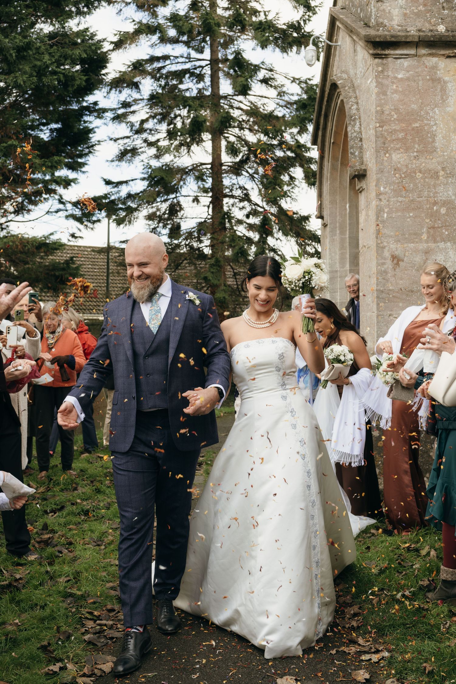 Happy newlyweds walk through shower of confetti outside historic stone church as guests celebrate.
