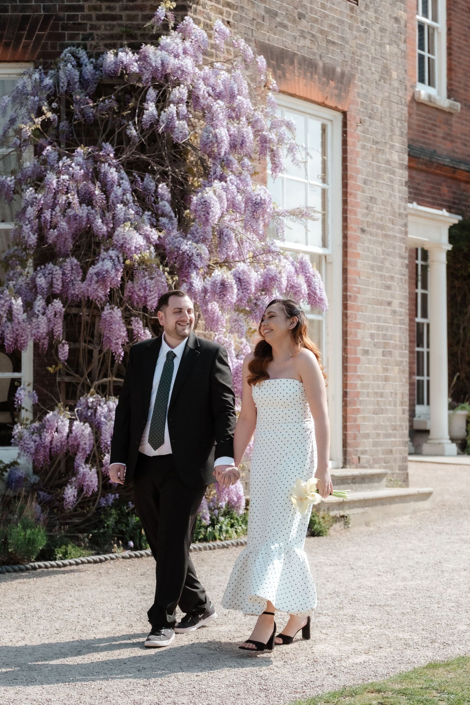Bride and groom walk hand-in-hand outside a brick manor, framed by blooming purple wisteria on a sunny day.