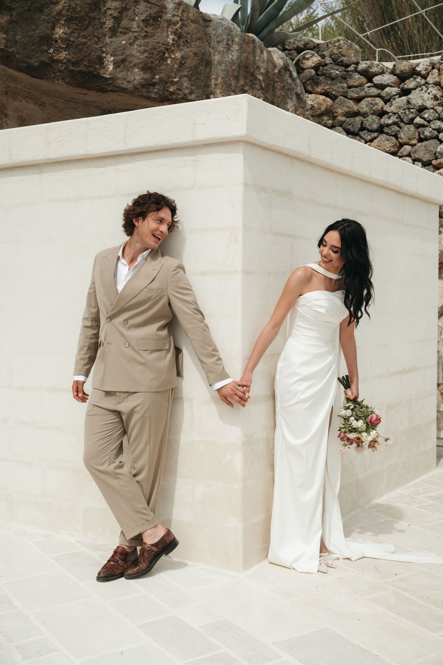 Bride in white gown and groom in beige suit hold hands against a white stone wall at an outdoor wedding venue.