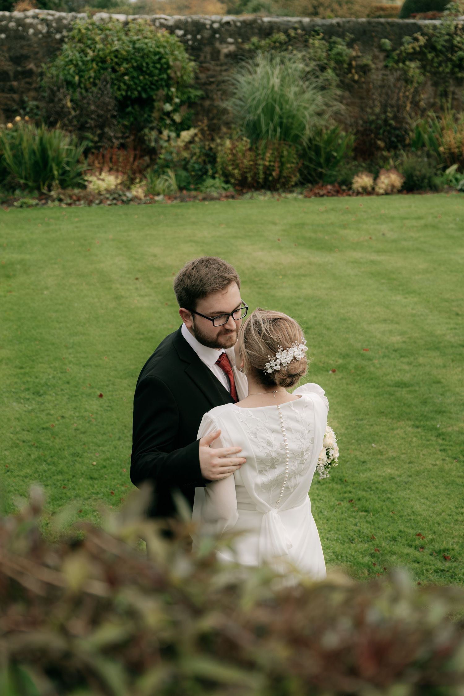 Intimate wedding couple embrace on manicured lawn with garden wall backdrop.