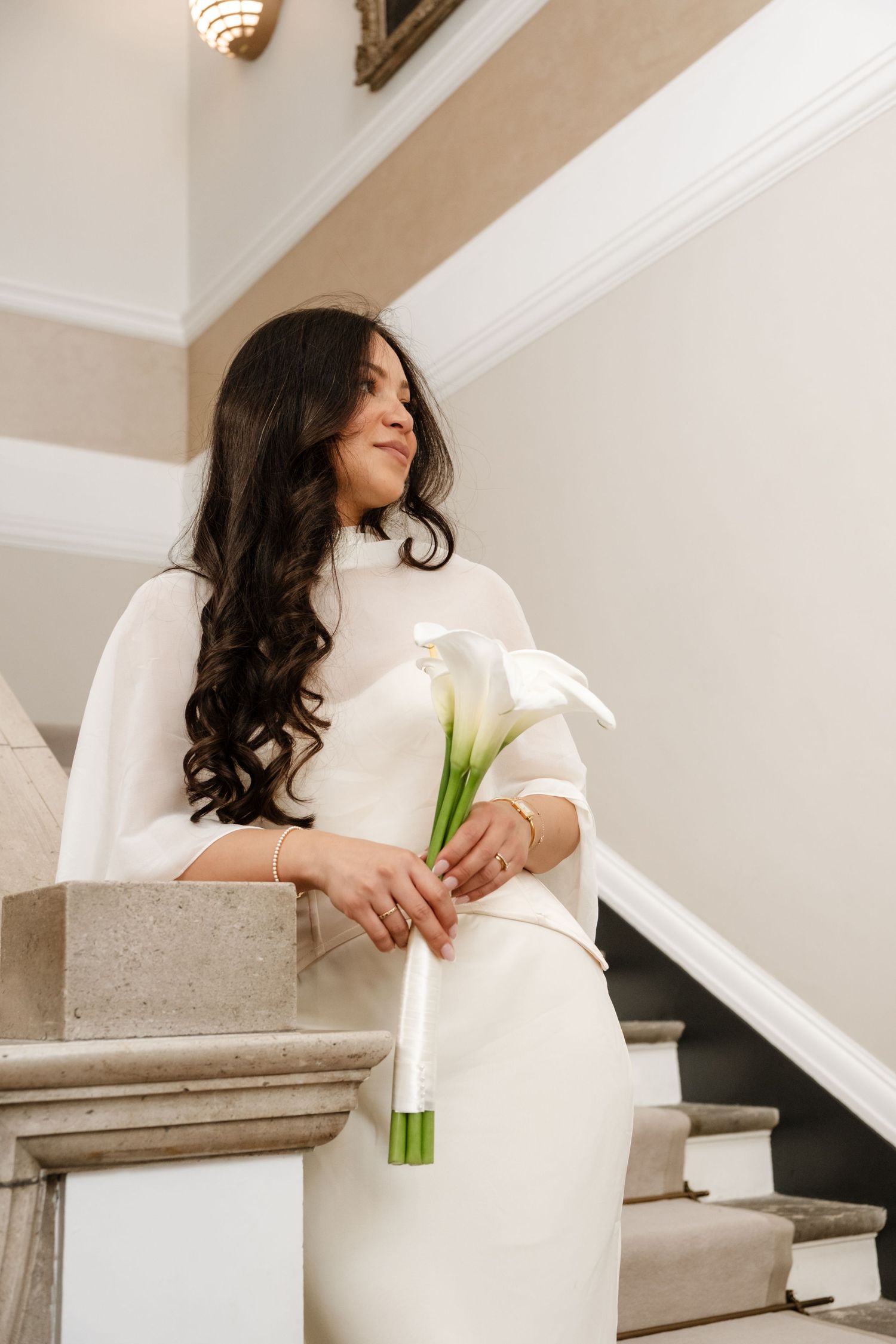 Elegant bride in white cape dress holding calla lily bouquet on marble staircase, smiling softly.