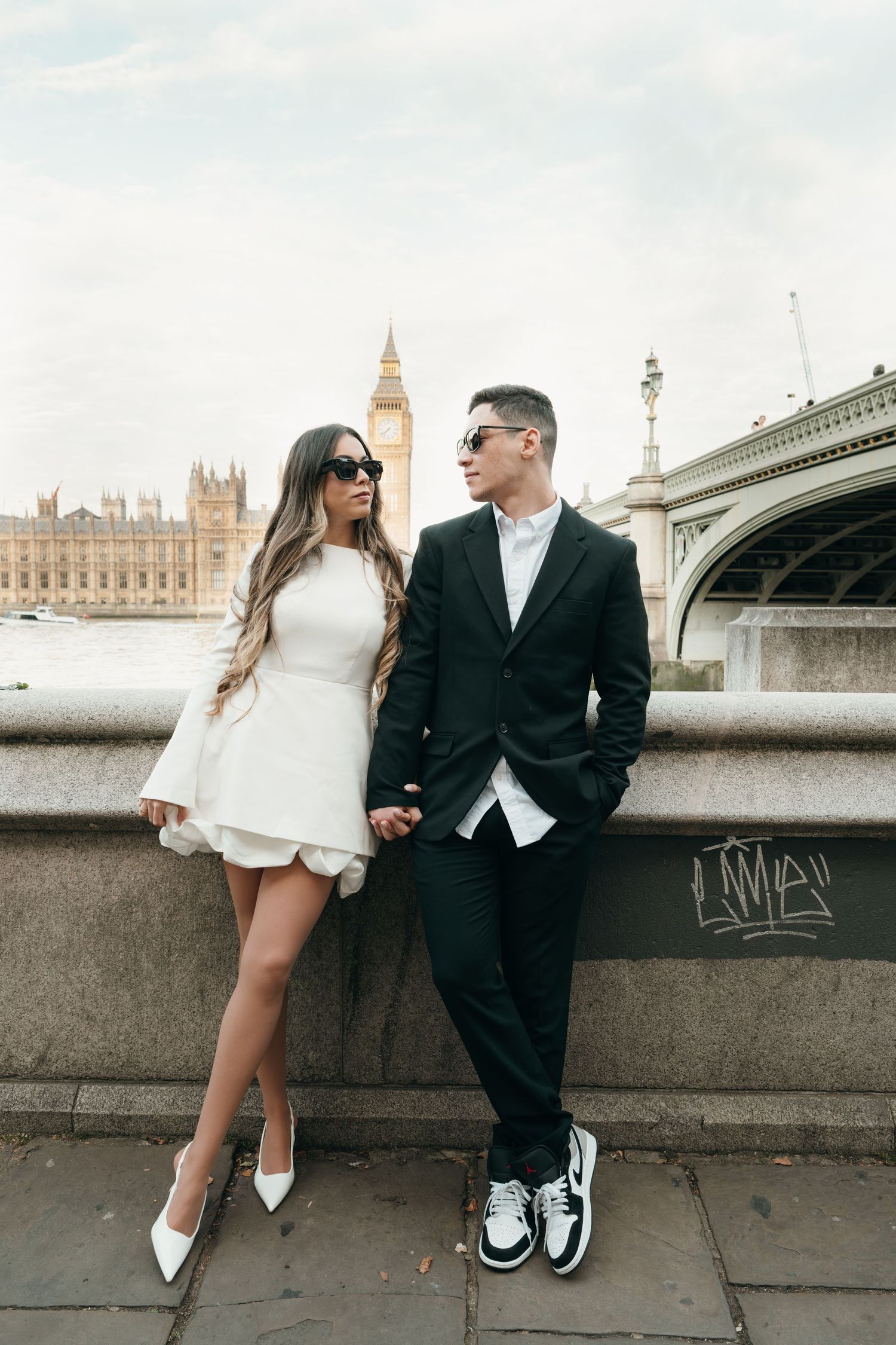 A stylish couple poses along the Thames River embankment with Westminster Palace in view.