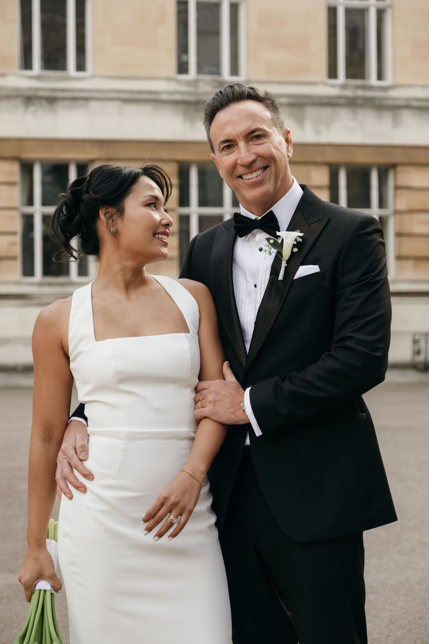 Elegant couple in wedding attire smiling together outside classic stone building.