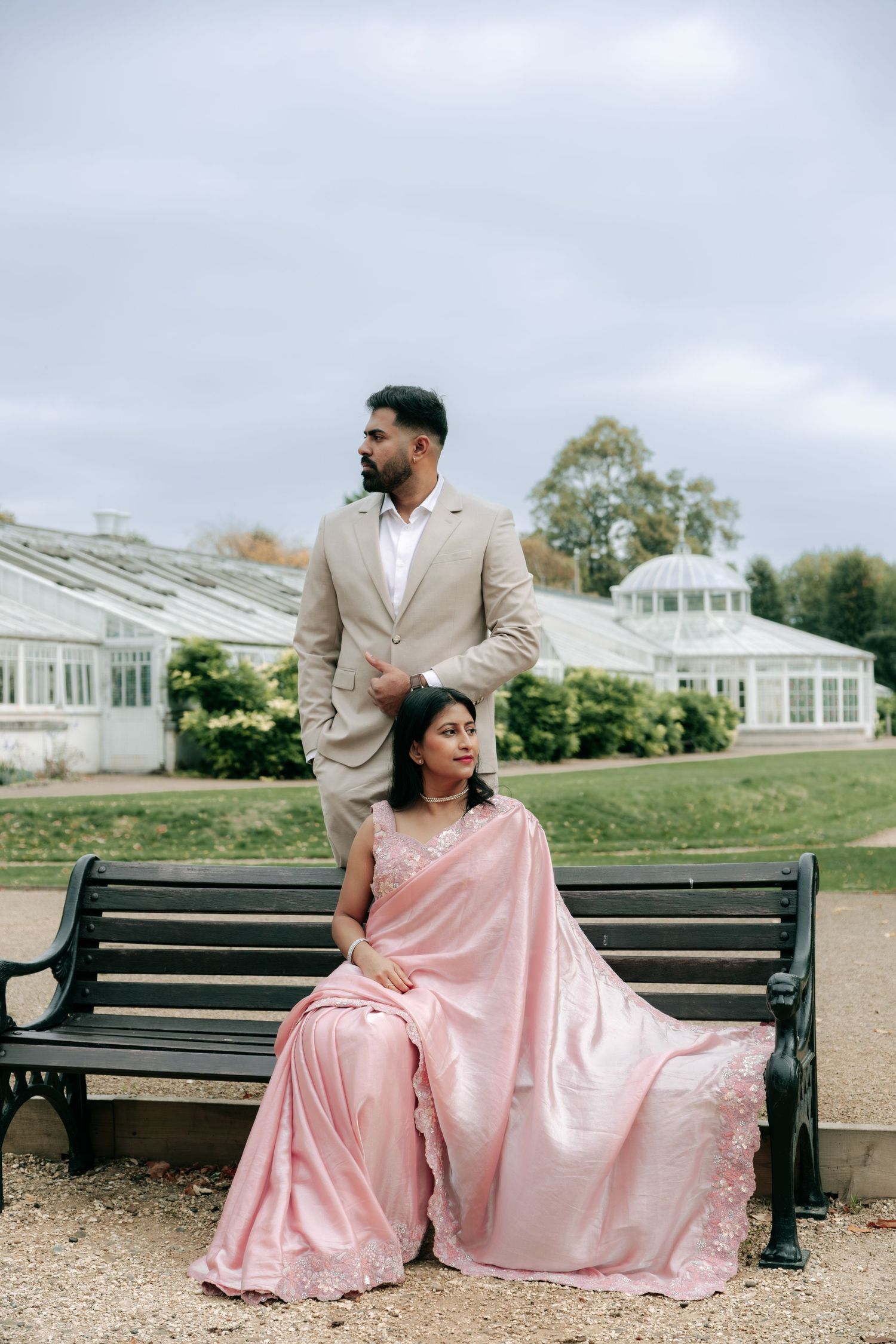 Elegant couple in formal wear poses on bench near greenhouse in traditional South Asian clothing.