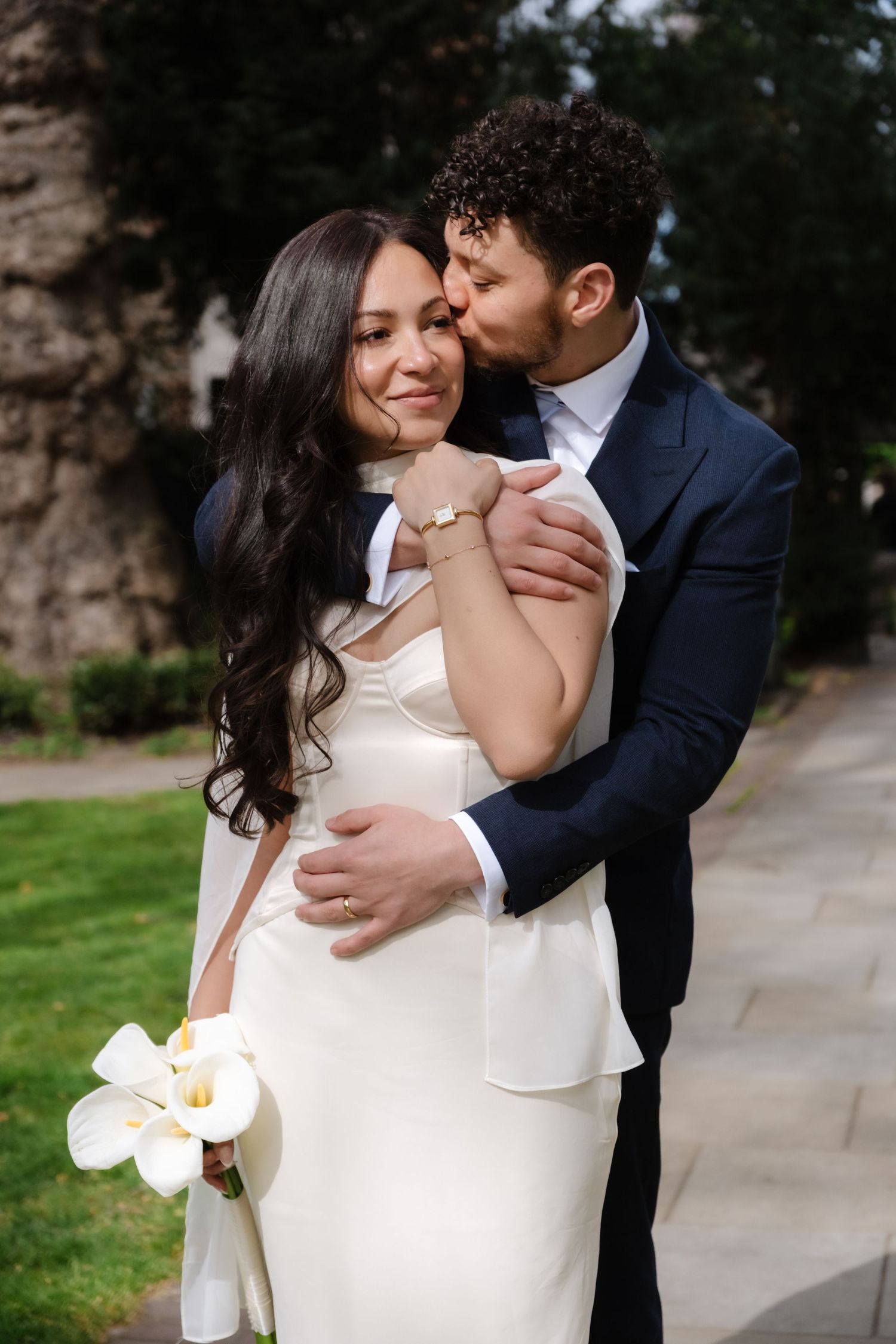 Groom in navy suit kisses bride in white gown holding calla lilies outdoors on their wedding day.